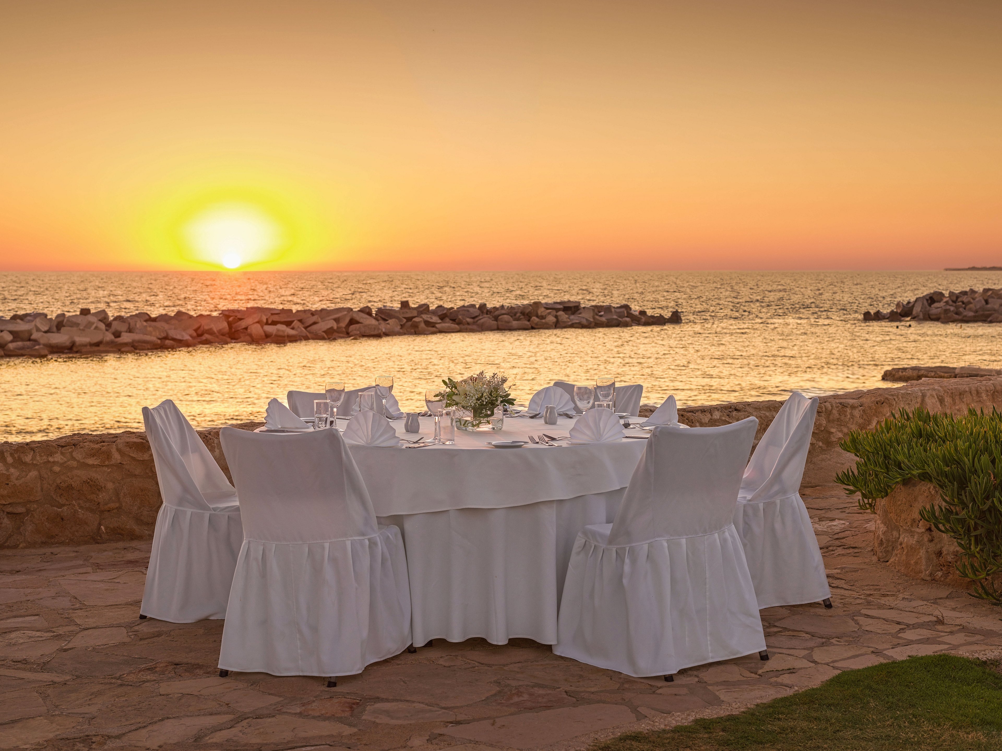 A round dining table covered in white linens is set for a formal meal beside the sea at sunset. The chairs are draped in matching white covers, and glassware and tableware are neatly arranged around a central floral centerpiece. The scene overlooks calm water and a rocky breakwater, with the sun low on the horizon casting a golden glow. Warm orange and yellow tones fill the sky, creating a romantic and tranquil coastal atmosphere.