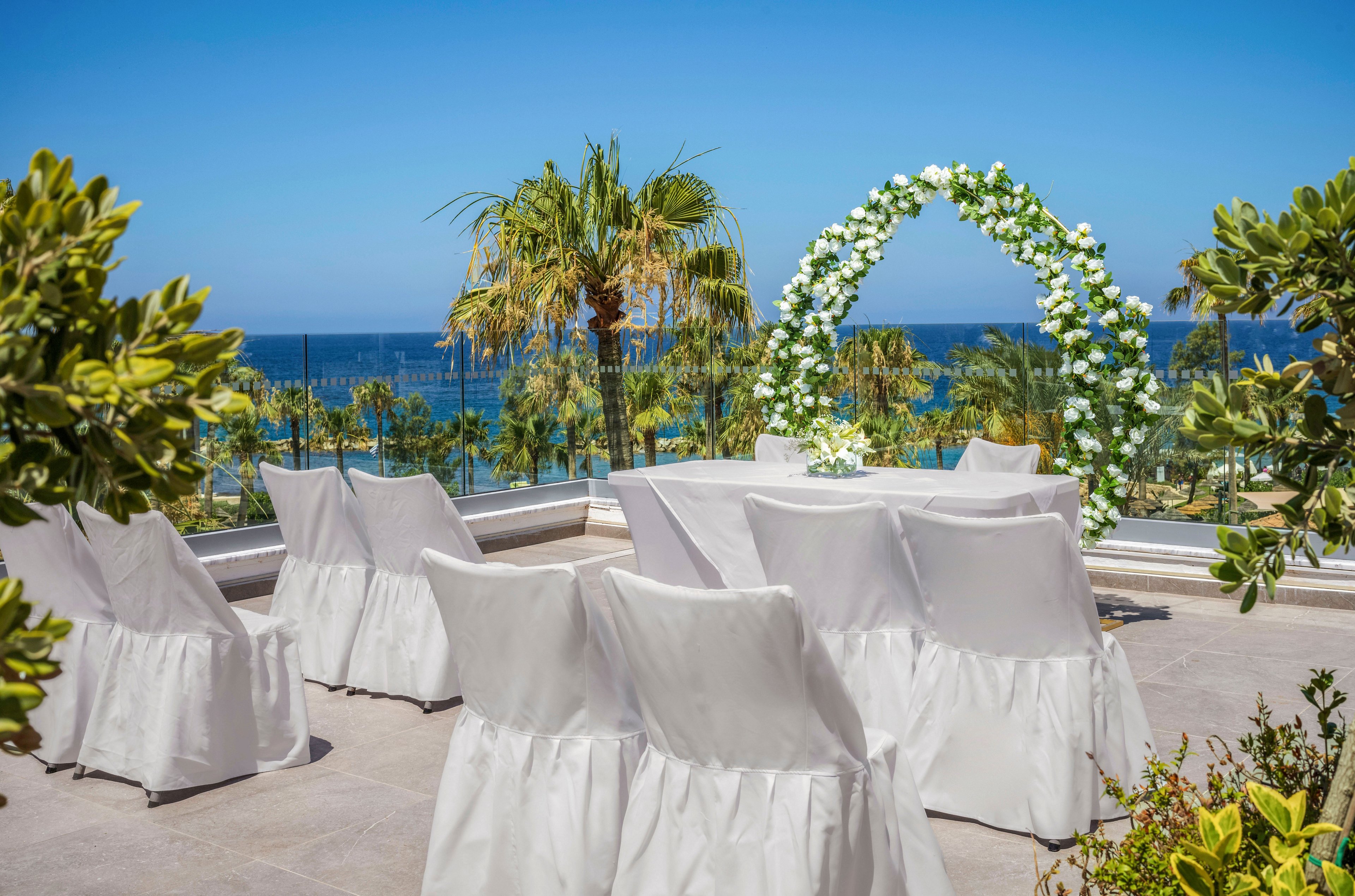 An elegant outdoor wedding ceremony setup overlooks a bright blue sea. White-covered chairs are arranged facing a floral arch decorated with white flowers and greenery. Palm trees and lush plants frame the terrace, adding a tropical feel to the scene. The clear sky and vibrant colors create a sunny, romantic atmosphere by the water.