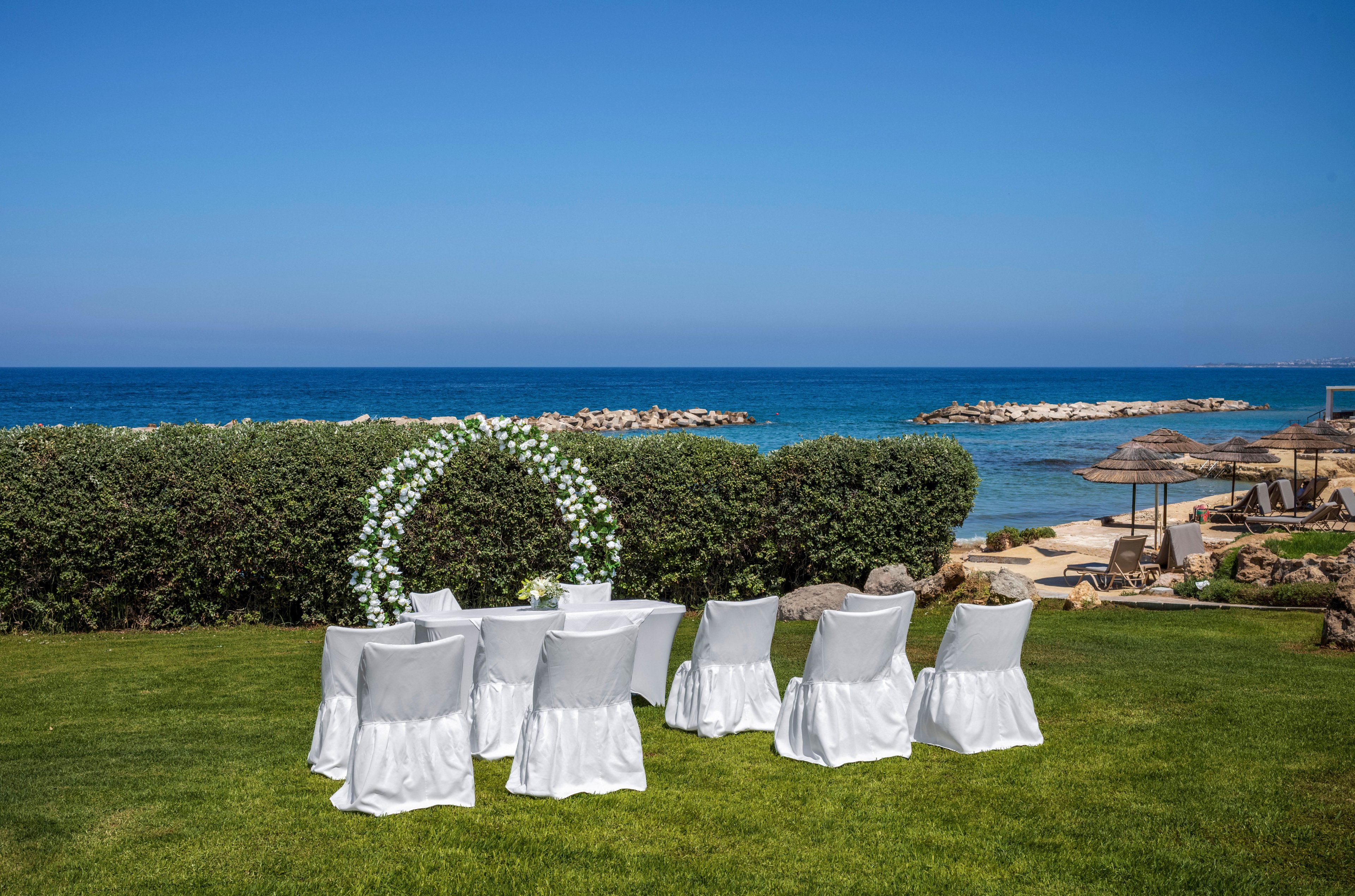 A small wedding ceremony setup is arranged on a green lawn overlooking the sea. White chairs with covers are placed in rows facing a floral arch. The arch is decorated with white flowers and greenery, framed by a hedge and a clear blue sky. In the background, rocky breakwaters and beach umbrellas line the shoreline, creating a tranquil coastal atmosphere.