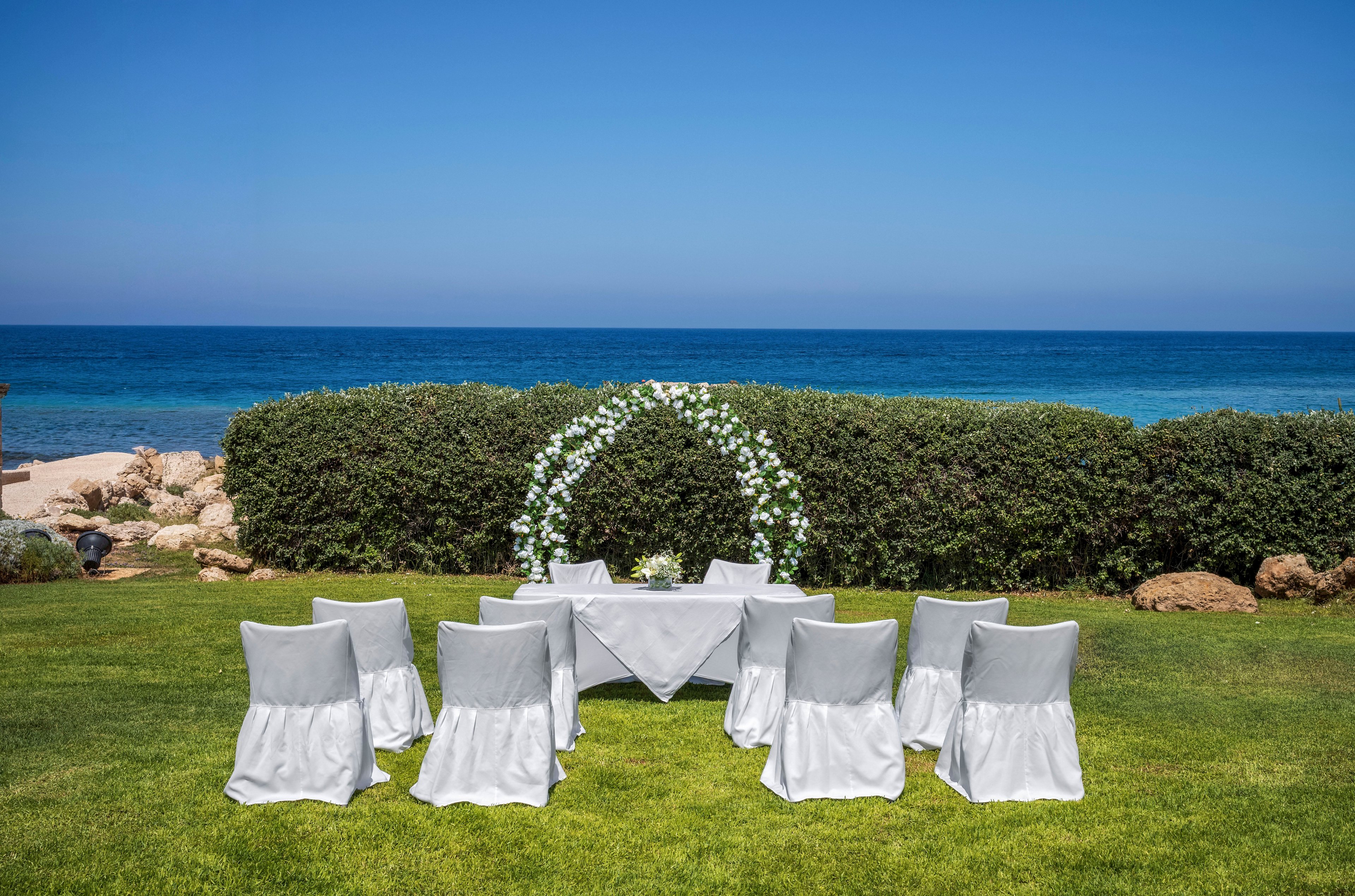 An intimate wedding ceremony setup is arranged on a lush green lawn overlooking the sea. White-covered chairs surround a small table draped in white cloth, positioned in front of a floral arch. The arch is decorated with white flowers and greenery, standing against a backdrop of trimmed hedges. Beyond the hedges, the blue ocean and clear sky create a serene and romantic atmosphere.