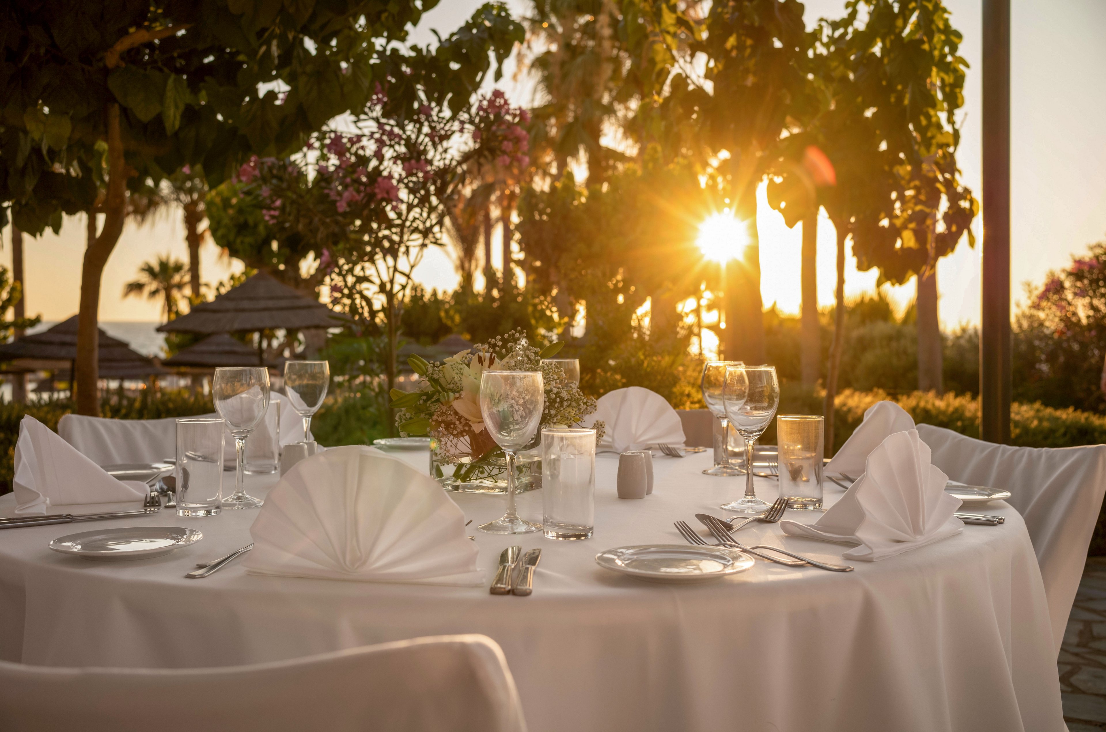 A round, white-clothed dinner table is set outdoors with neatly folded napkins, glassware, and cutlery. The scene is framed by lush greenery, palm trees, and distant thatched structures, suggesting a resort or garden setting. Warm sunlight filters through the trees, creating a golden glow over the table and surroundings. The overall atmosphere feels romantic and sophisticated, ideal for a special event or celebration.