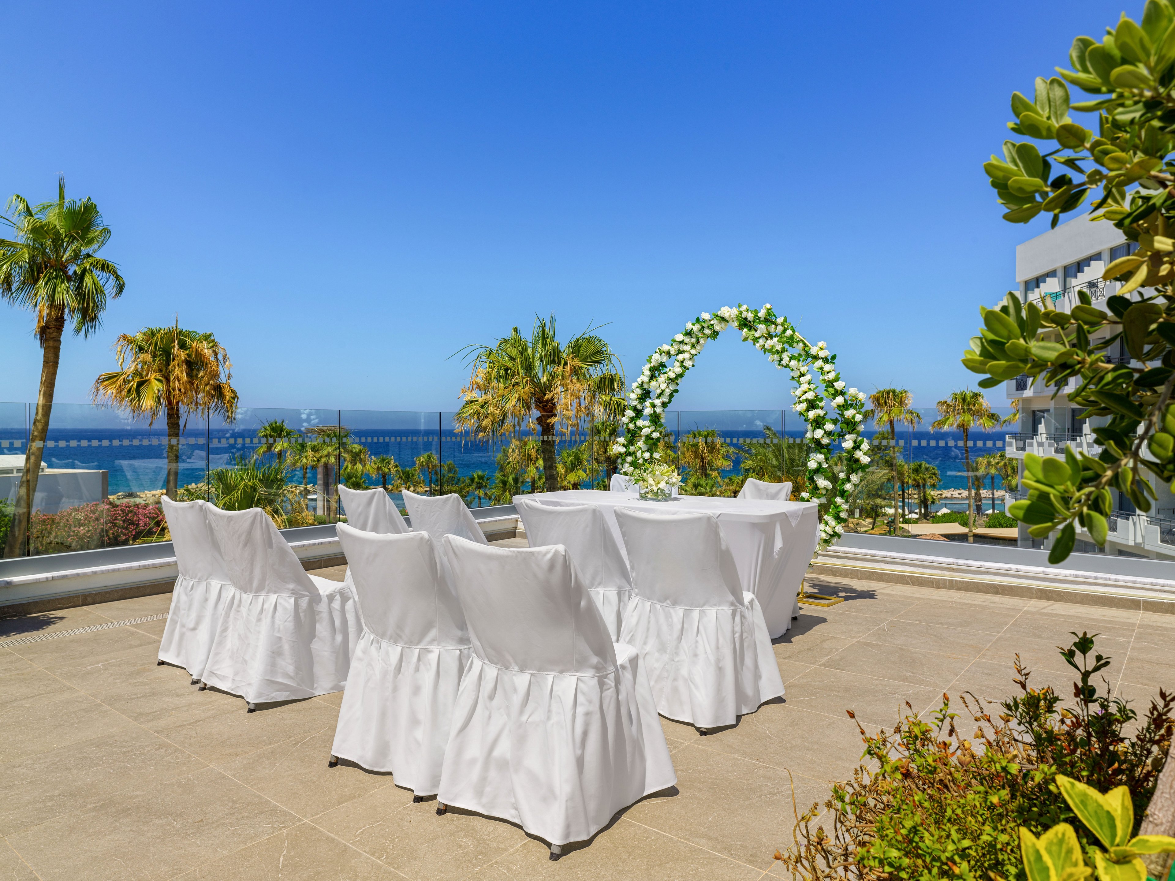 An elegant outdoor wedding ceremony setup overlooks a bright blue sea. White-covered chairs and a table are arranged on a terrace facing a floral arch. Palm trees and modern buildings frame the view under a clear sunny sky. The scene suggests a peaceful, romantic celebration in a coastal resort setting.
