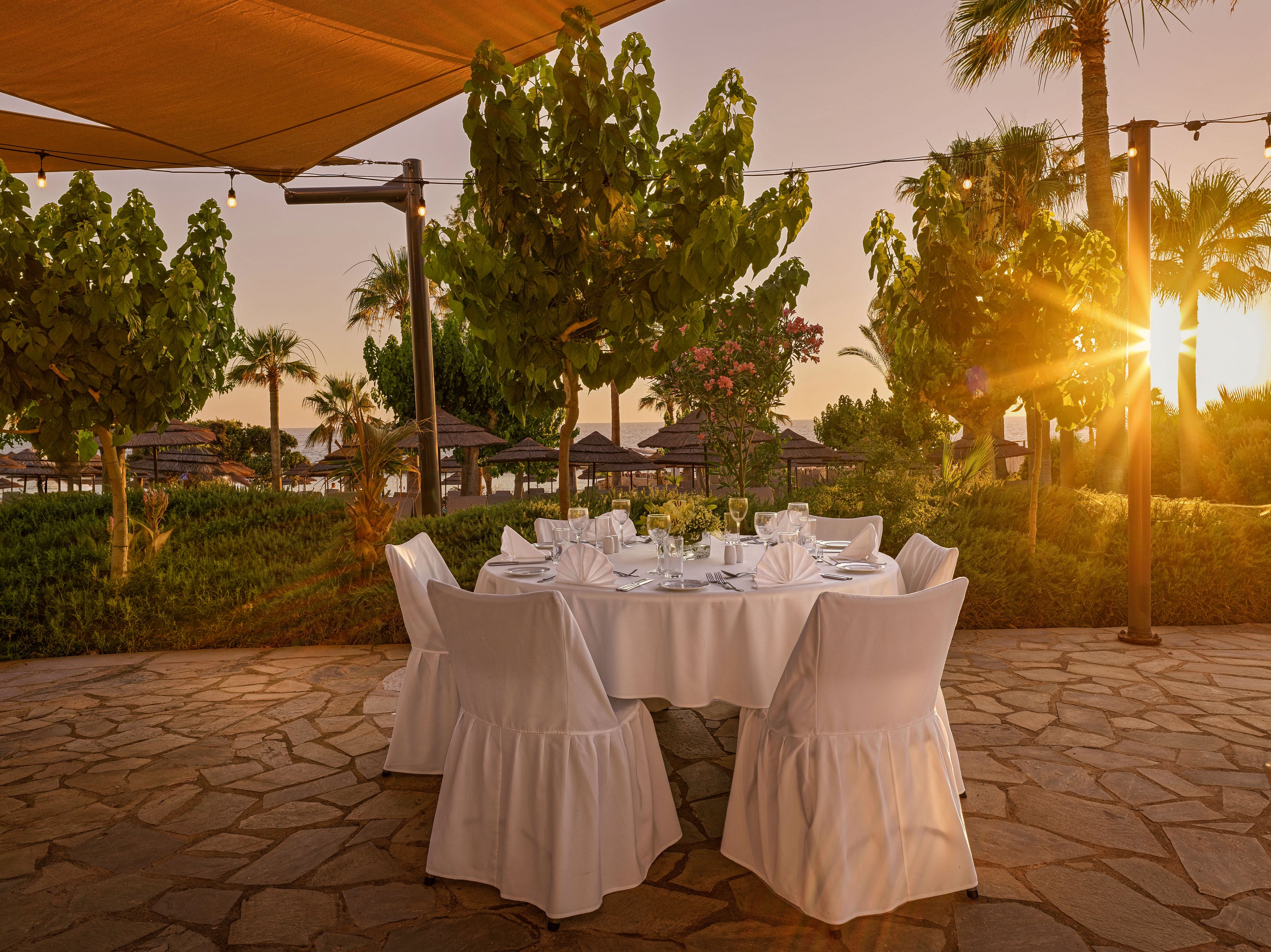 A round dining table is set outdoors with white cloth and chair covers, prepared for a formal meal. The scene is surrounded by lush green trees and landscaped gardens under a warm evening sky. String lights and a sunshade canopy add ambiance as the sun sets behind palm trees. The stone patio surface and neatly arranged tableware create a refined, inviting atmosphere.
