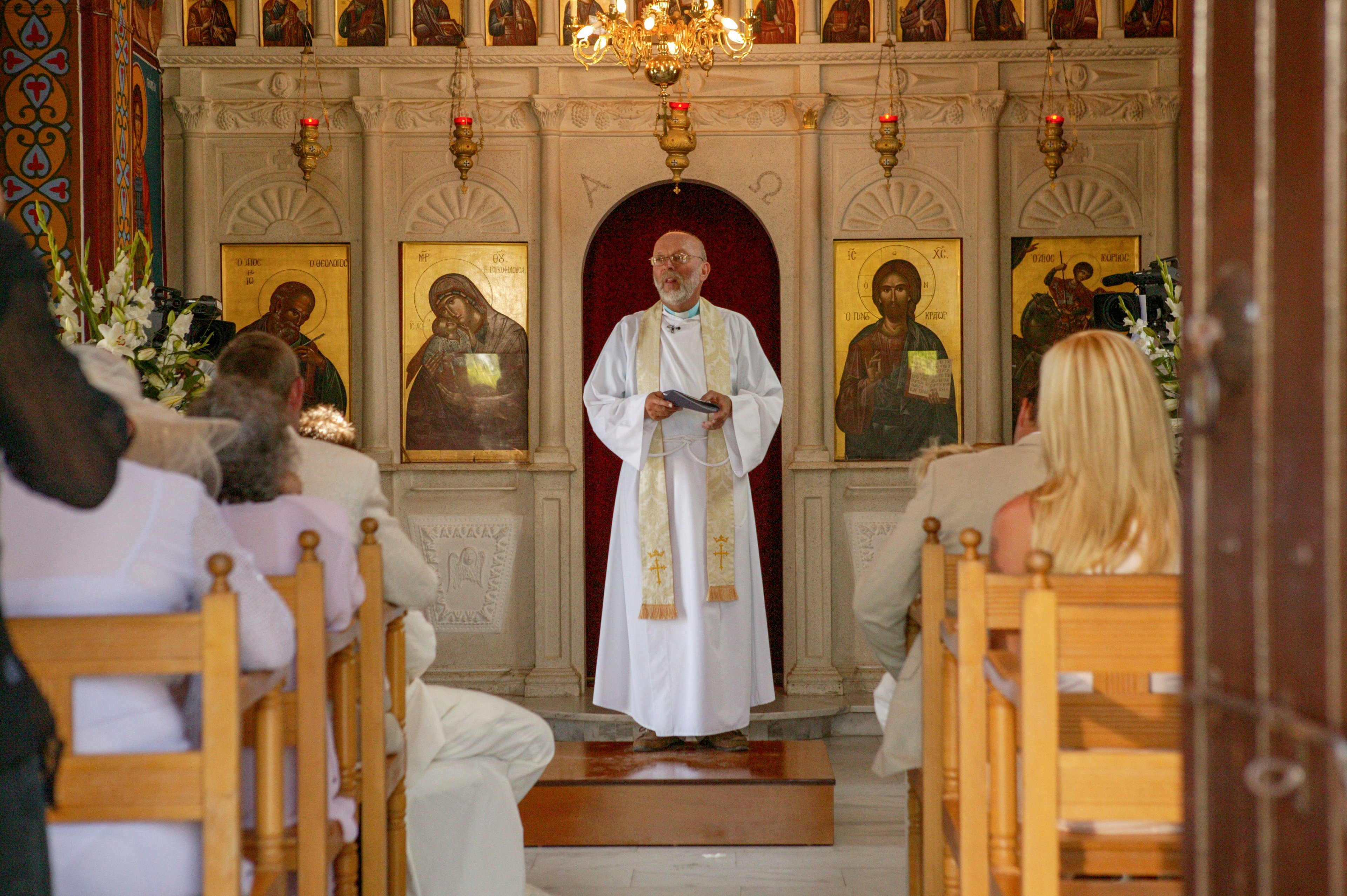 A priest in white vestments stands at the front of a small church, addressing seated attendees. The interior features an ornate iconostasis with religious paintings and gold accents behind him. Wooden pews are filled with people dressed in light-colored formal clothing, suggesting a special ceremony or service. Warm lighting from a chandelier above adds a reverent and intimate atmosphere to the scene.