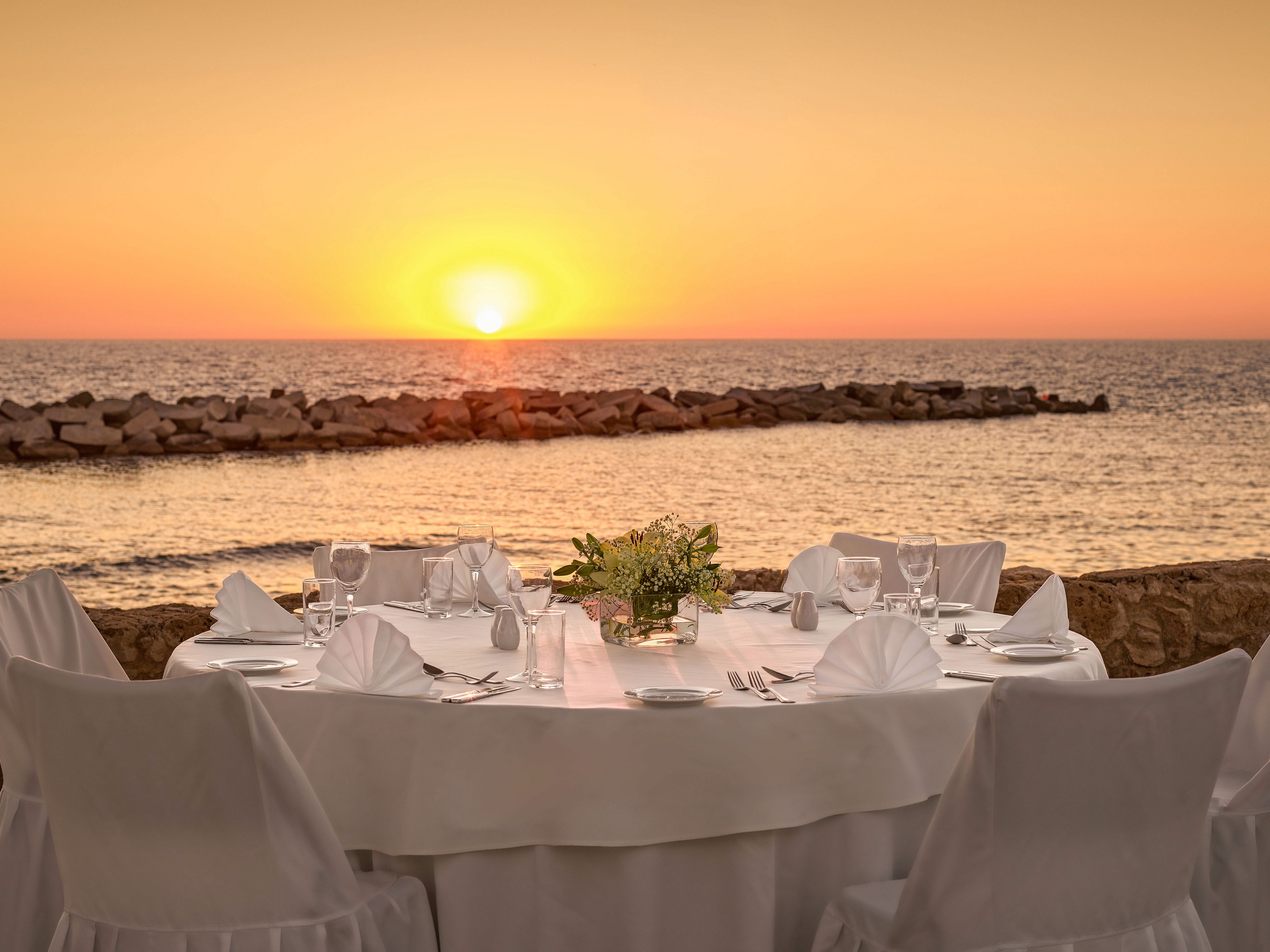 A round dining table is set elegantly with white linens and folded napkins overlooking the sea at sunset. Glassware, plates, and cutlery are neatly arranged, suggesting a formal meal or celebration. The sun is low on the horizon, casting warm golden light across the calm water and rocky breakwater. The overall scene feels romantic and tranquil, ideal for an evening event by the ocean.