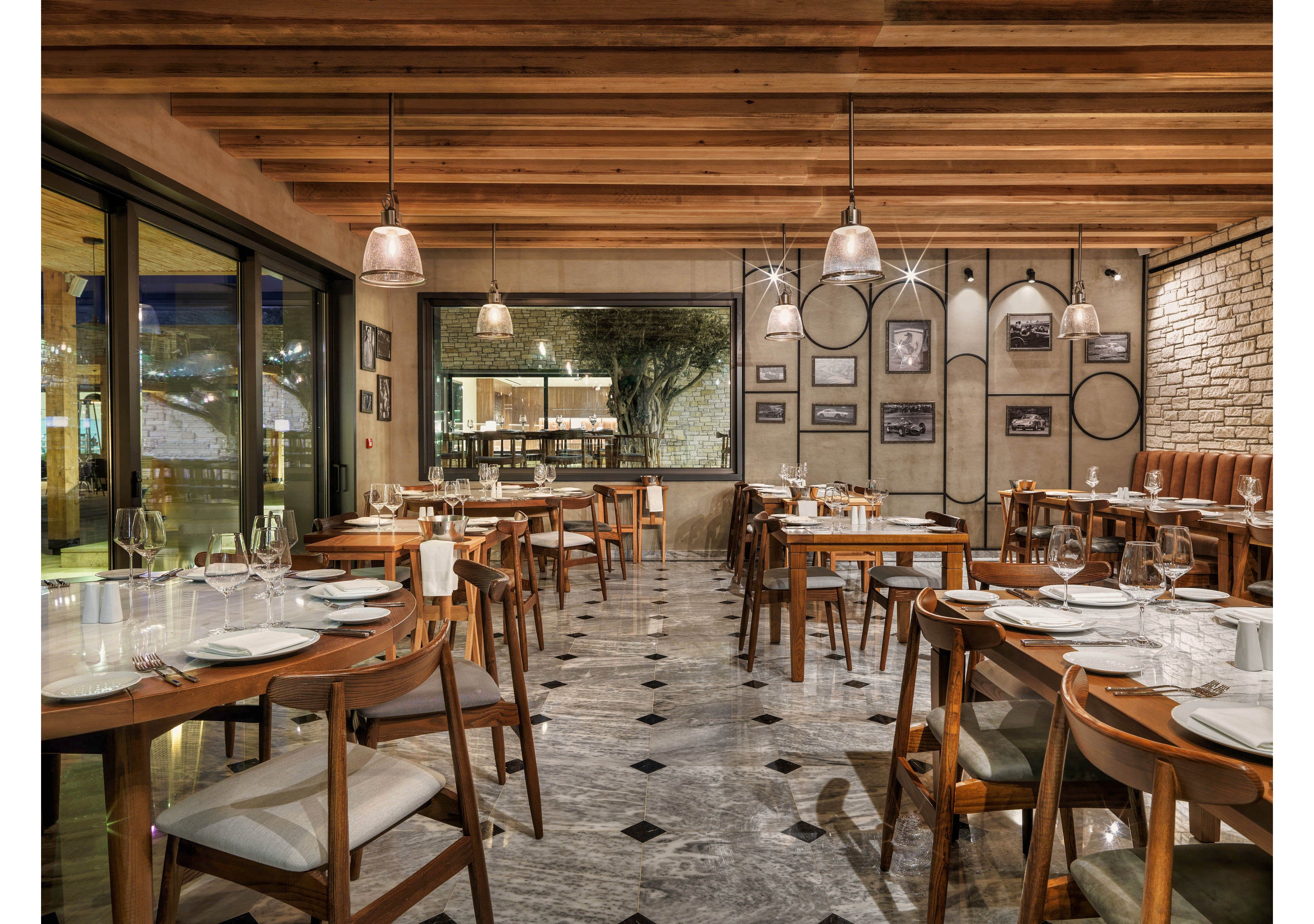 Contemporary restaurant interior with neatly arranged wooden tables and chairs is shown empty and ready for guests. The setting features a polished stone floor with dark diamond inlays and a ceiling of exposed wooden beams. Pendant lights hang over the tables, illuminating white plates, glassware, and folded napkins. Large windows and framed wall art add depth and character to the cozy dining space.