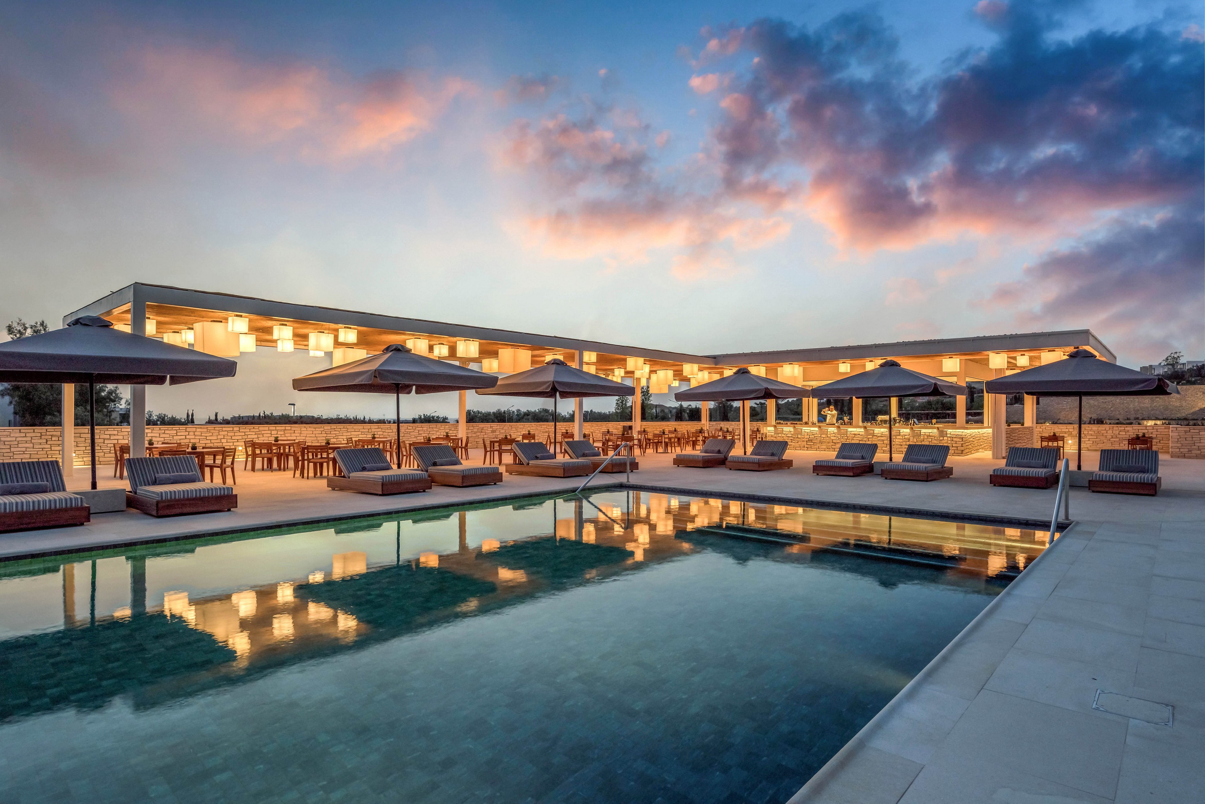 A contemporary resort swimming pool is shown at dusk with calm water reflecting the illuminated building. Several lounge chairs and large umbrellas line the poolside terrace, creating a relaxing leisure area. Warm artificial lights contrast with the cool blue and pink tones of the evening sky. The scene suggests a tranquil vacation setting with outdoor dining and relaxation spaces.