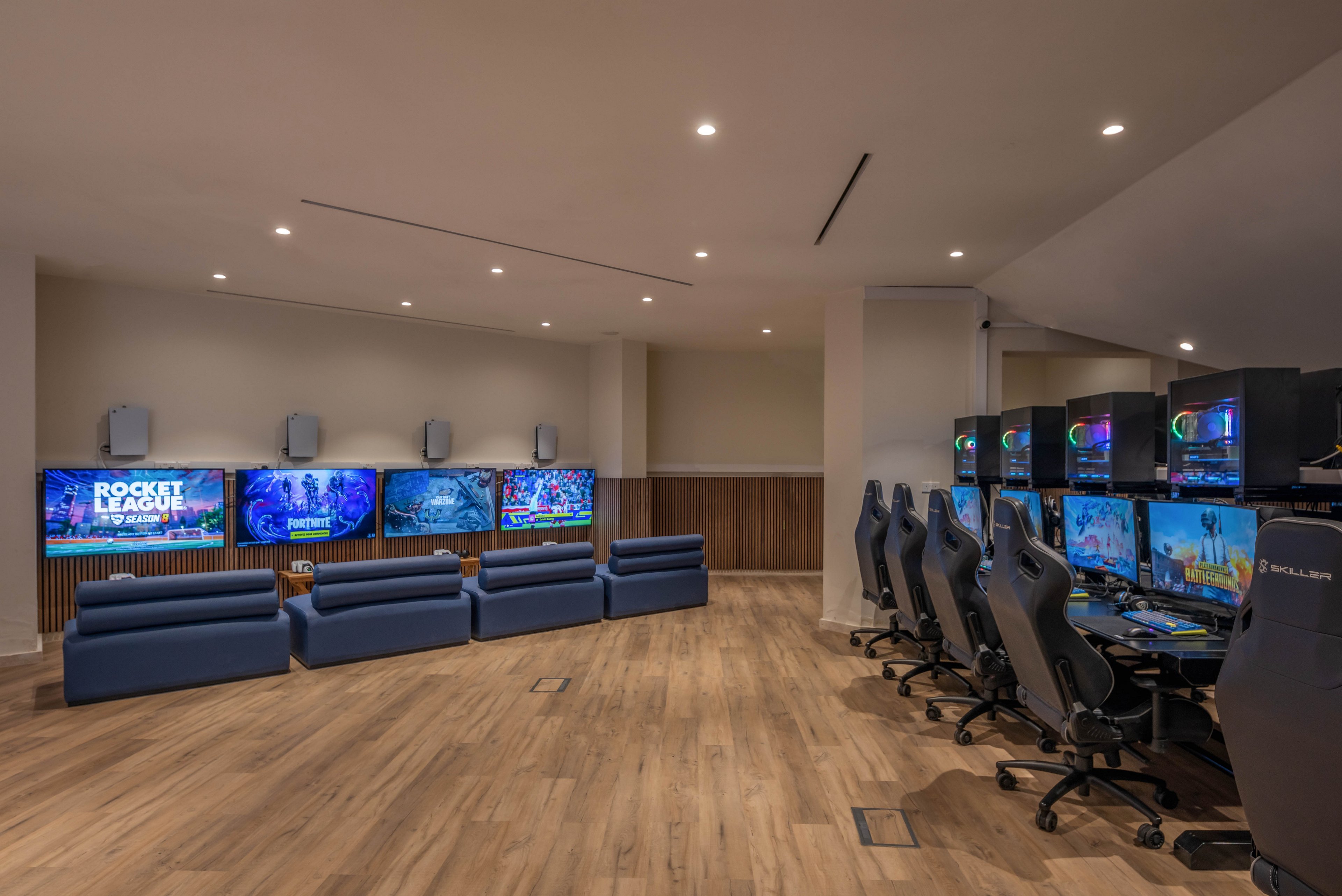 A spacious gaming lounge features multiple large wall-mounted screens displaying colorful video games. Comfortable blue sofas face the televisions on one side of the room, while a row of high-backed gaming chairs and desktop computers lines the opposite wall. Recessed ceiling lights and warm wood flooring create a clean, contemporary atmosphere. The room appears designed for group gaming sessions, esports, or social entertainment.