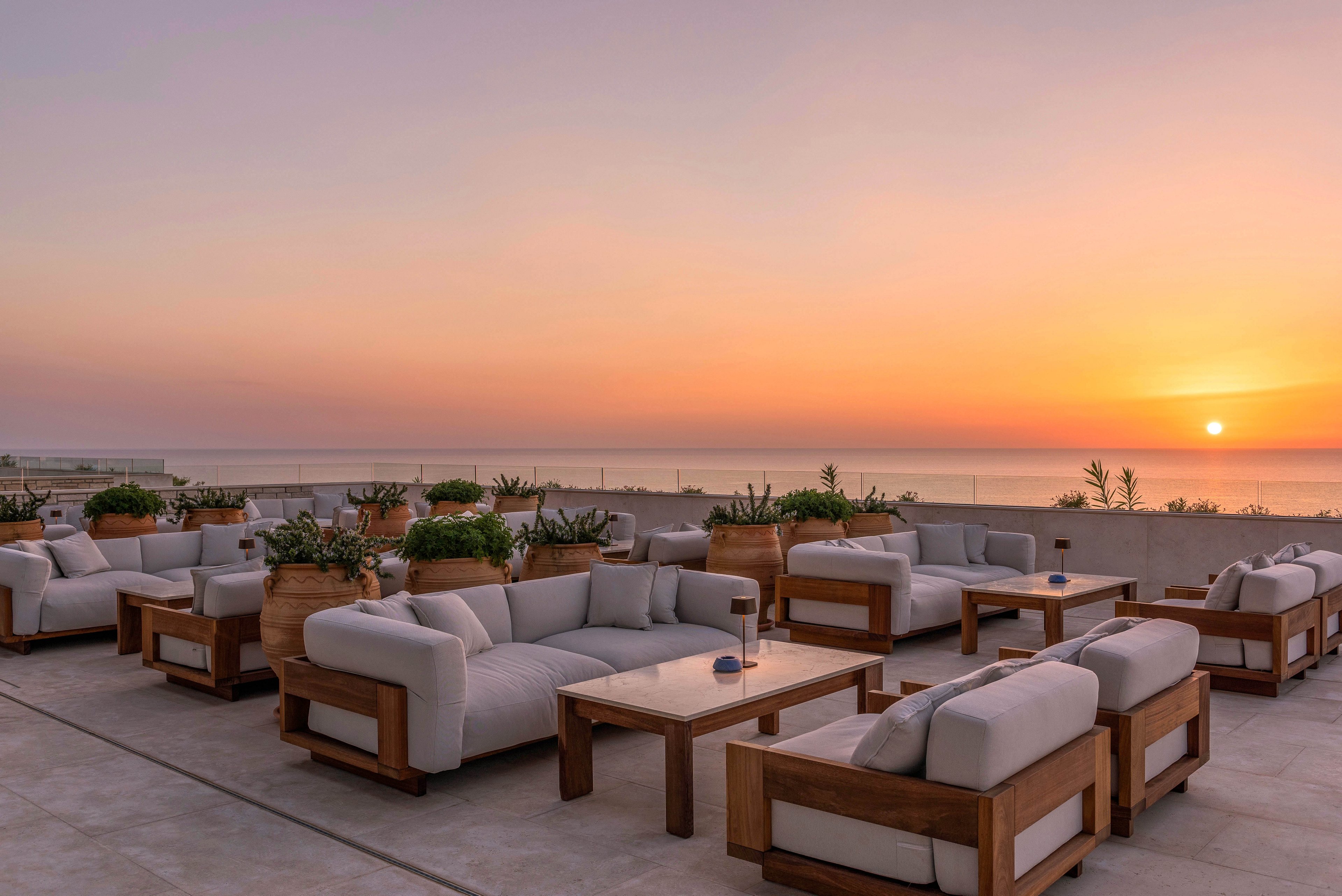 An elegant rooftop lounge features modern wooden sofas with light gray cushions arranged in cozy seating groups. Large planters with greenery are placed between the furniture, adding a natural touch to the contemporary terrace. The scene overlooks a calm body of water as the sun sets near the horizon, casting warm orange and purple tones across the sky. Soft ambient lighting begins to glow on the tables, enhancing the relaxed, upscale atmosphere.
