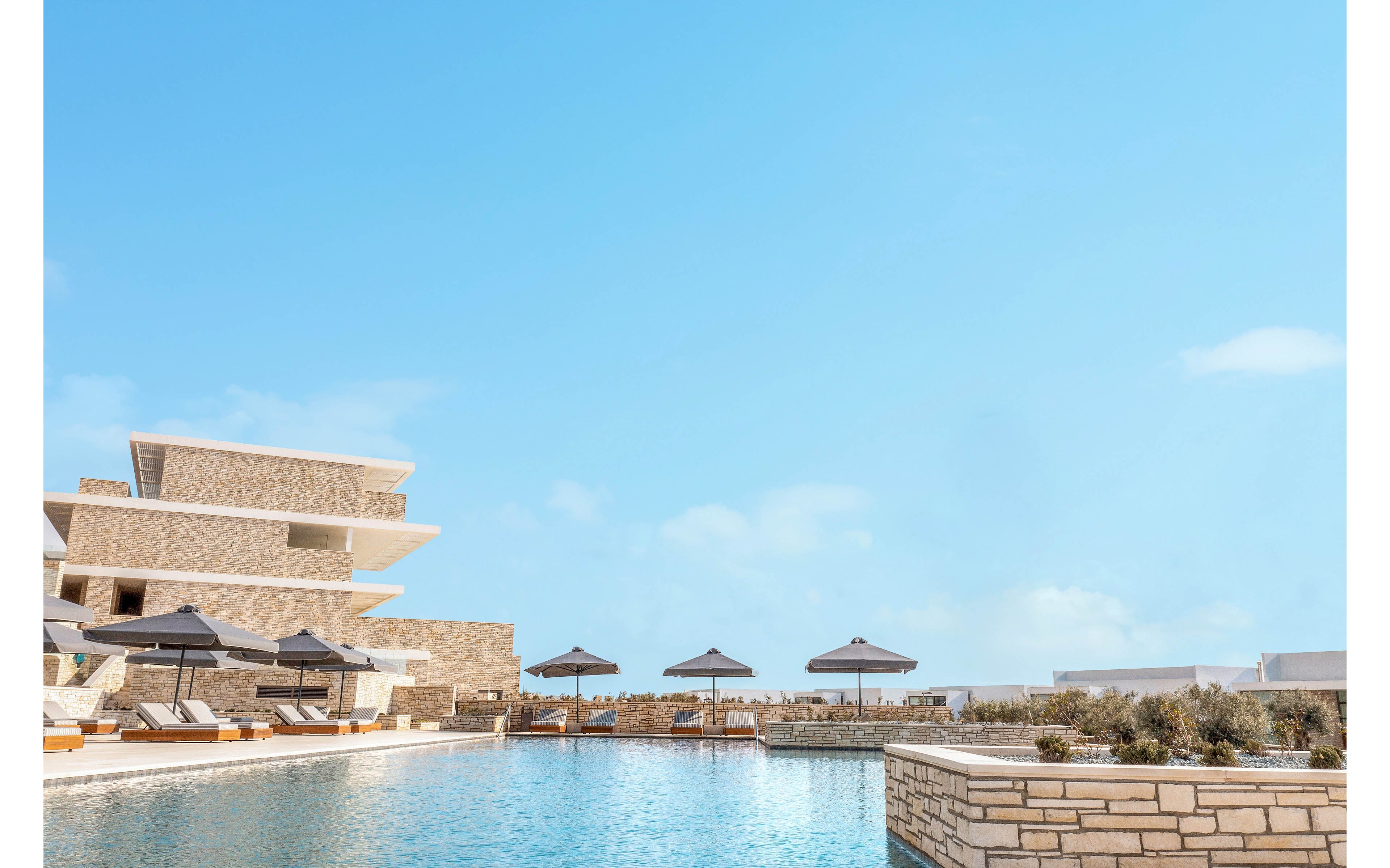 A woman in a white one-piece swimsuit stands on the edge of a modern outdoor swimming pool. The setting appears to be a luxury resort with contemporary stone architecture and layered building structures. Sun loungers and dark umbrellas line the poolside, suggesting a relaxing vacation atmosphere. The bright daylight and clear blue sky create a clean, minimalist and serene mood.