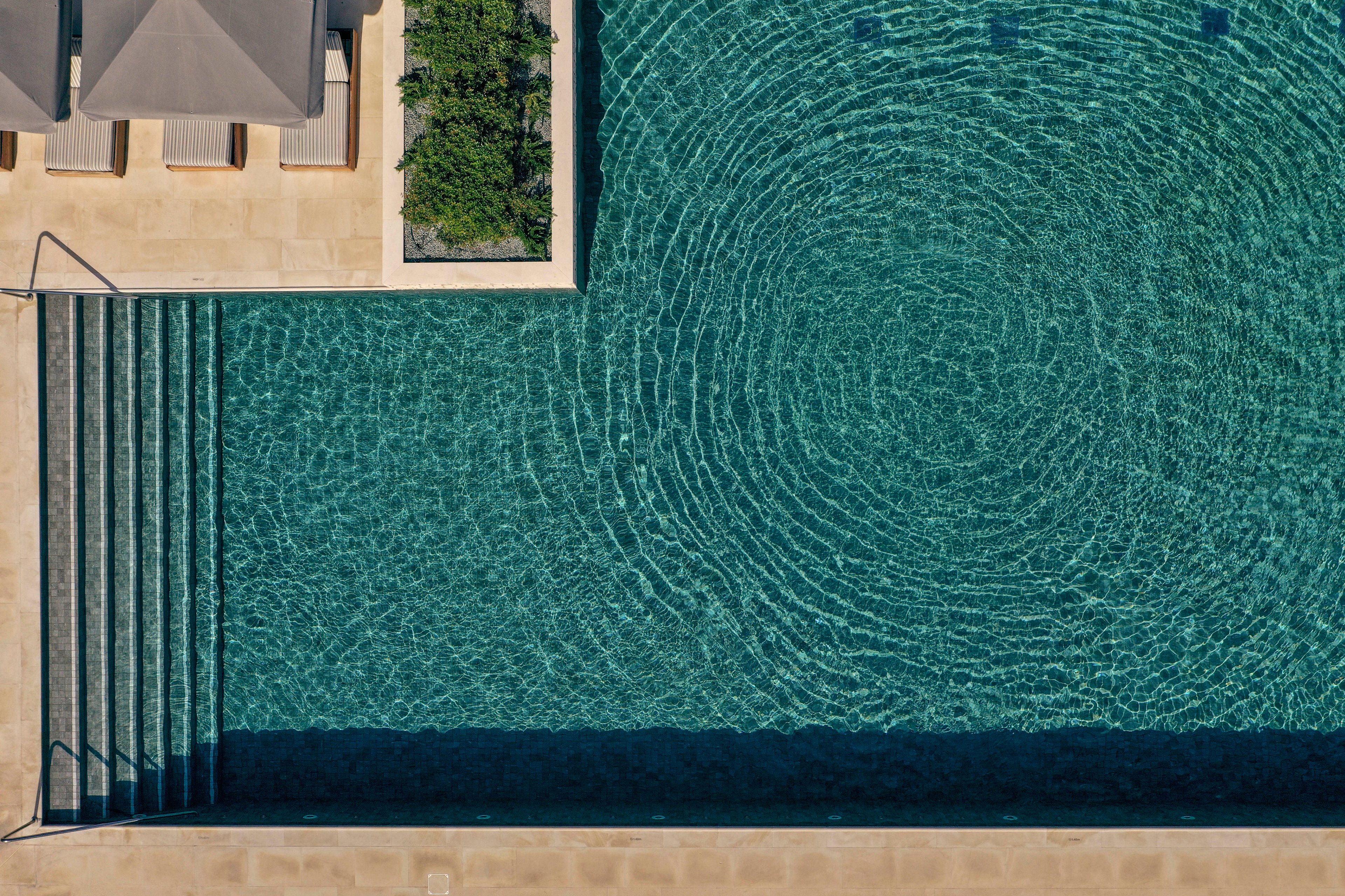 An aerial view shows a lone swimmer floating in the center of a large rectangular swimming pool. Concentric ripples spread outward across the clear blue-green water, creating a striking pattern. Wide stone steps and surrounding beige pool deck frame the scene with clean geometric lines. The overall atmosphere feels calm, minimal, and serene under bright daylight.