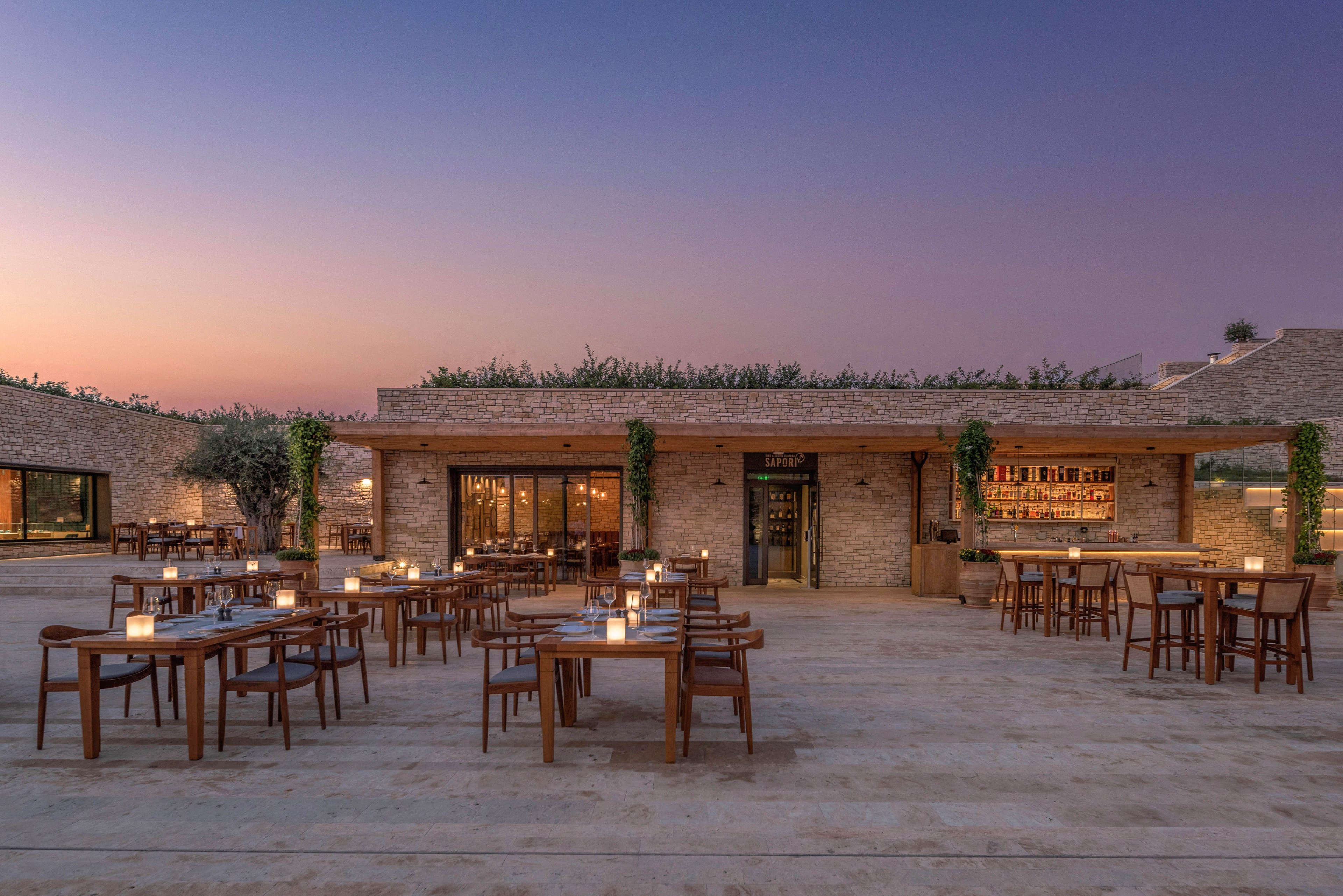 An elegant open-air restaurant terrace is shown at dusk, with wooden tables and chairs neatly arranged across a wide stone patio. Soft candlelight glows on each table, creating a warm and inviting atmosphere. The building features a modern rustic design with stone walls, large glass doors, and a bar area visible inside. The sky transitions from deep blue to soft orange, enhancing the tranquil evening ambiance.