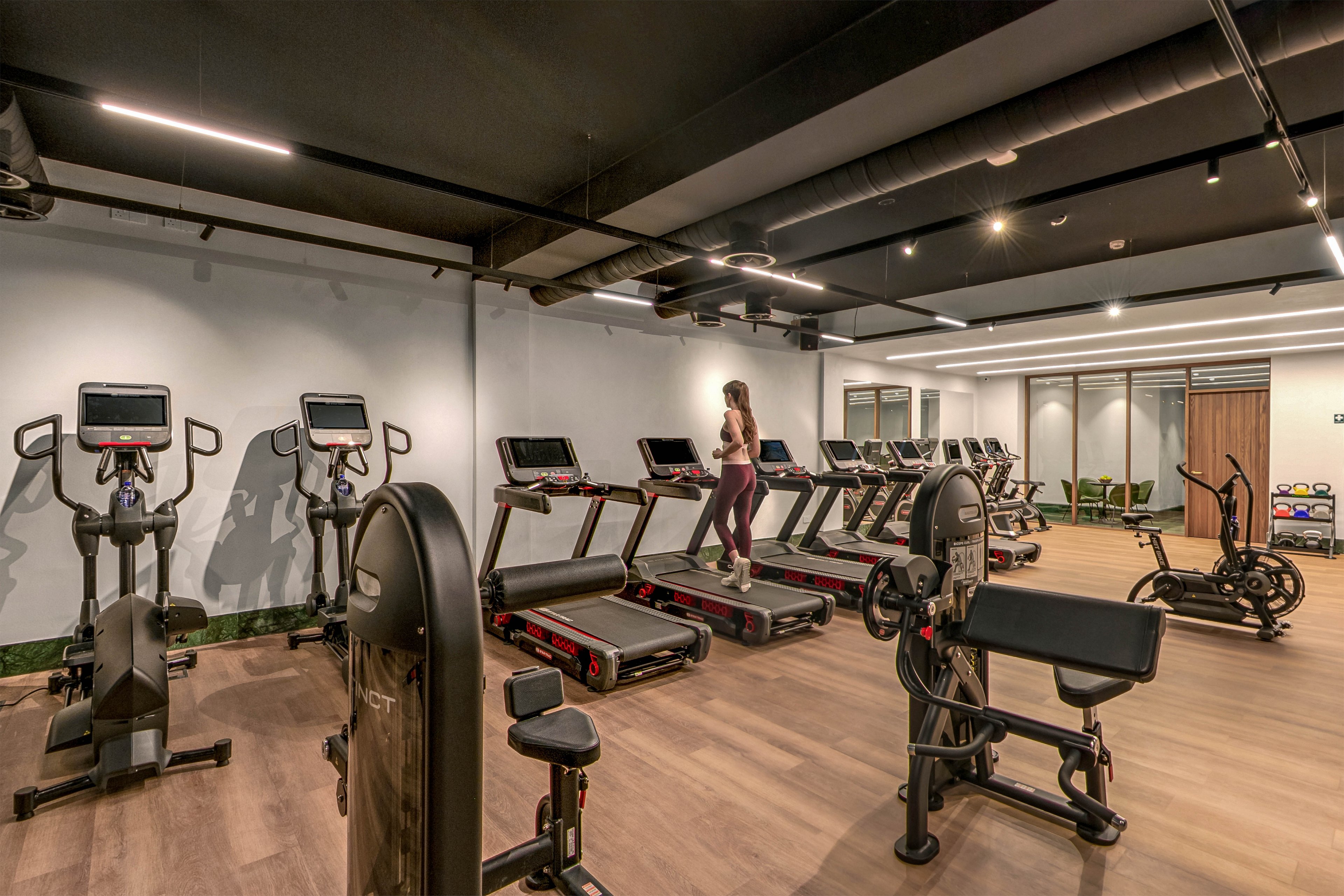 A spacious modern gym features rows of treadmills and exercise bikes on a wooden floor. A woman in athletic wear walks on a treadmill in the center of the room. Strength training machines and cardio equipment are neatly arranged under a dark industrial-style ceiling. Bright, even lighting and clean walls create a contemporary, professional fitness environment.