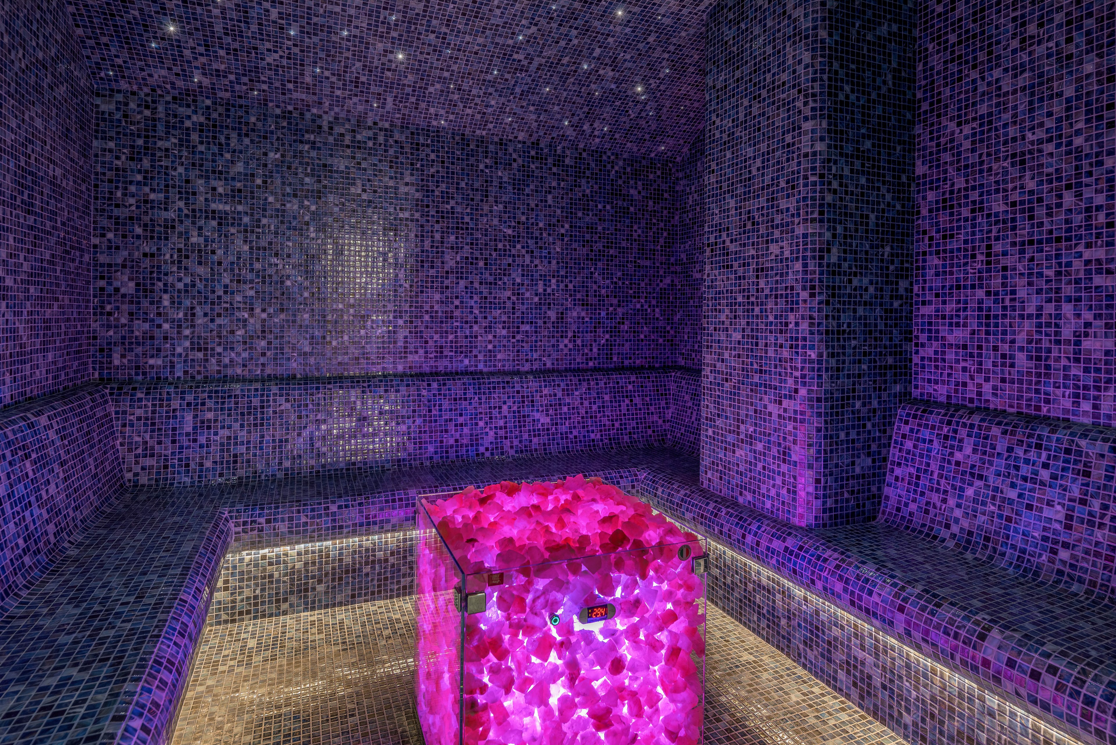 A contemporary steam room is fully lined with small mosaic tiles in shades of purple and blue. Built-in bench seating runs along the walls, creating a cozy, enclosed spa environment. At the center of the room, a glass cube filled with glowing pink crystals provides dramatic accent lighting. Recessed ceiling lights add to the ambient, luxurious atmosphere.