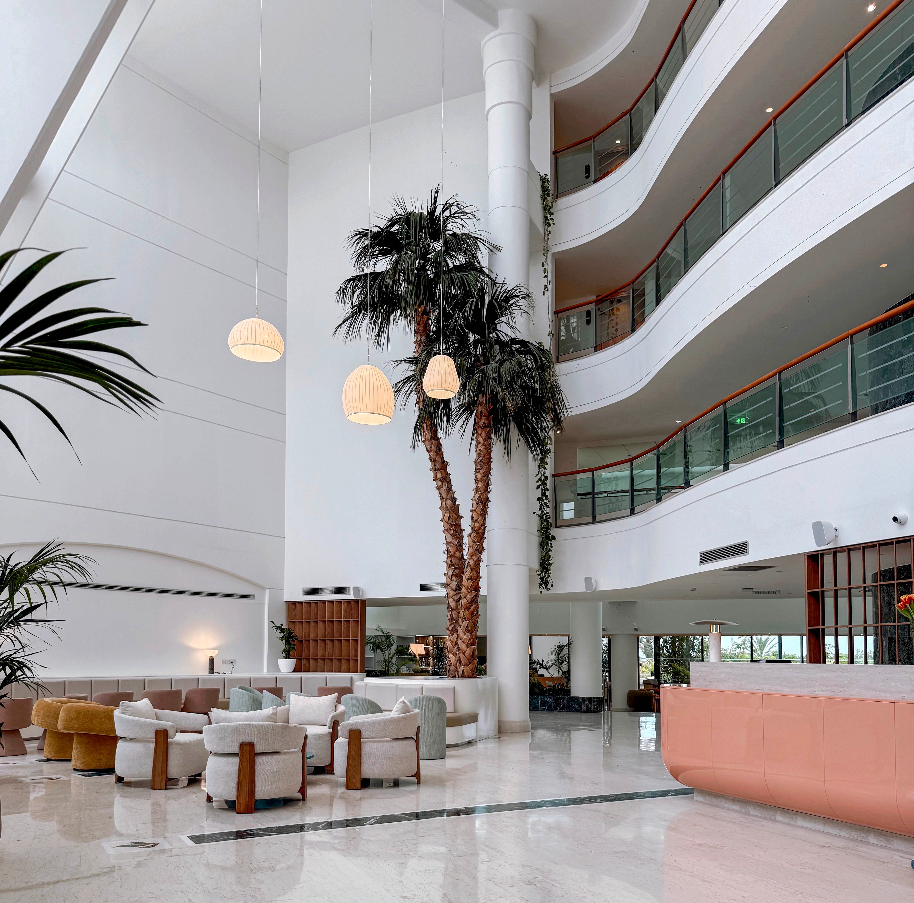 A spacious contemporary hotel lobby features tall indoor palm trees and a high white ceiling. Curved balconies with glass railings overlook the open atrium. In the foreground, stylish lounge seating in muted pastel tones surrounds low tables. Pendant lights hang above the seating area, adding warmth to the bright, minimalist interior. Large windows and glossy floors enhance the airy, modern atmosphere.