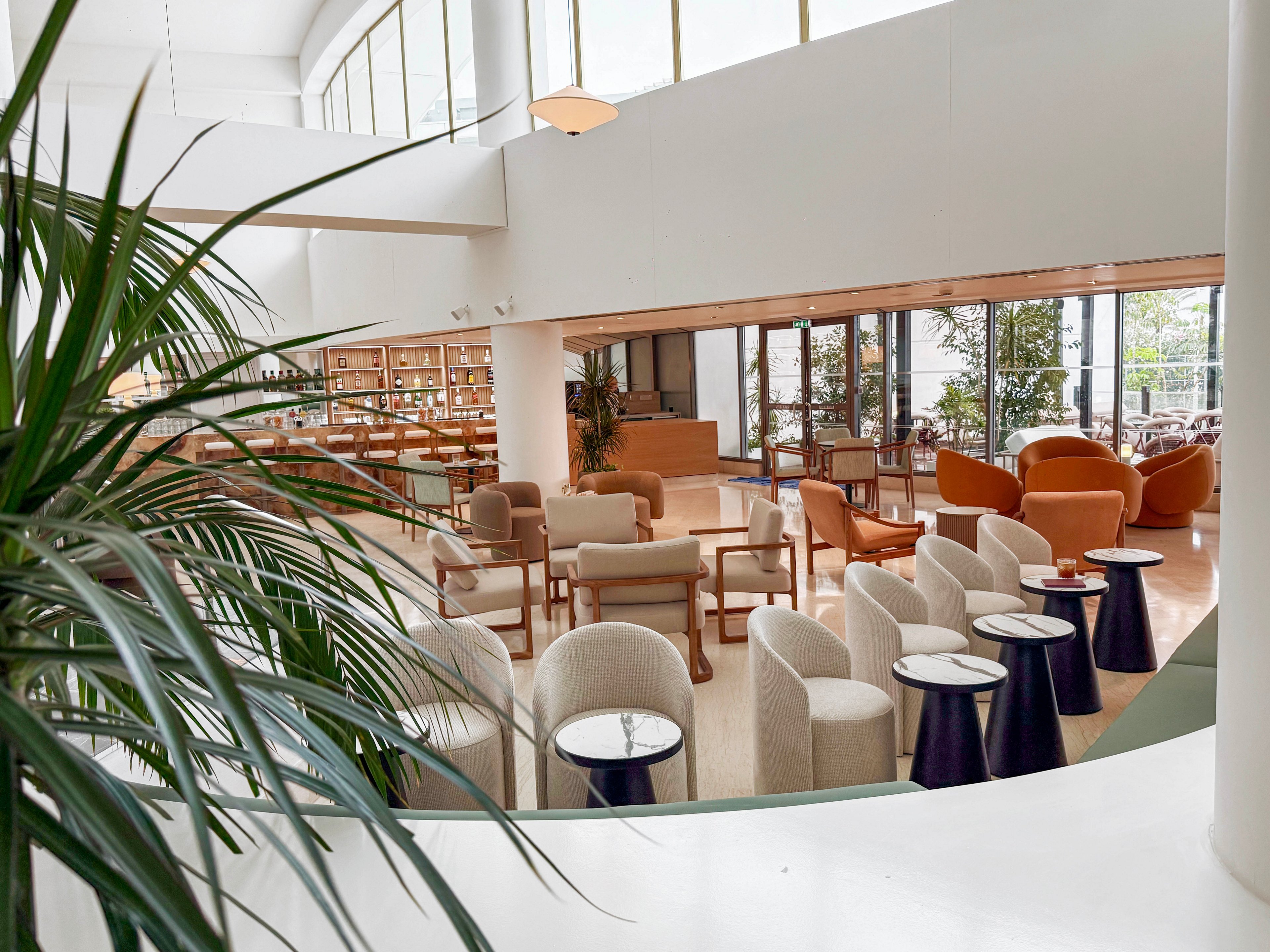 A spacious modern lounge features an array of contemporary chairs and small round tables arranged in clusters. Large floor-to-ceiling windows allow abundant natural light to fill the bright, open interior. Warm neutral tones and soft textures create a calm, inviting atmosphere. Potted plants and greenery add a touch of nature to the sleek architectural design.