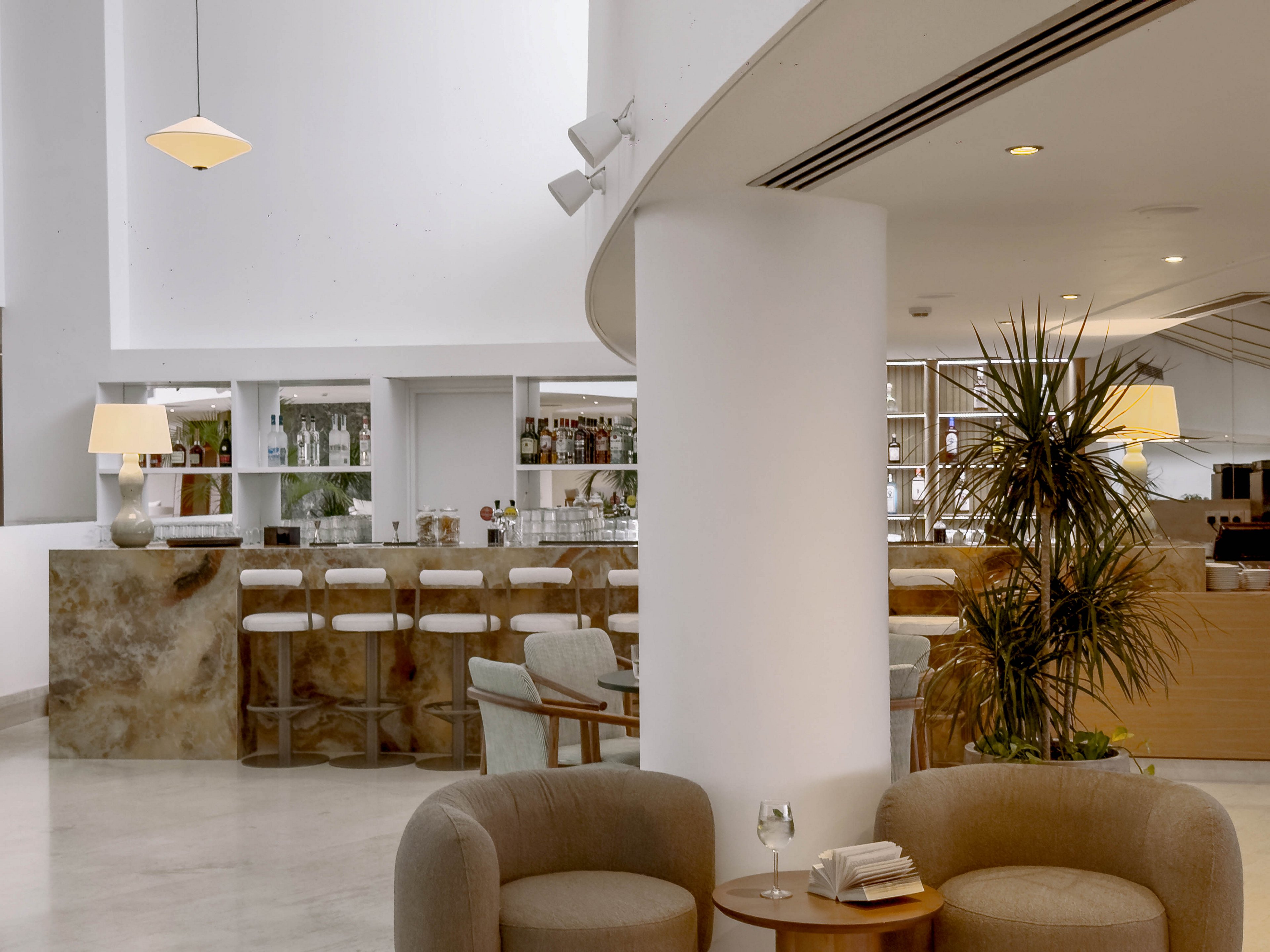 A contemporary hotel lounge features a sleek bar with high stools and a polished stone counter. Soft armchairs and a small table with drinks create an inviting seating area in the foreground. The space is bright and minimal, with white walls, warm lighting, and decorative plants adding a touch of greenery. Bottles and glassware are neatly arranged on shelves behind the bar, emphasizing a refined, upscale atmosphere.