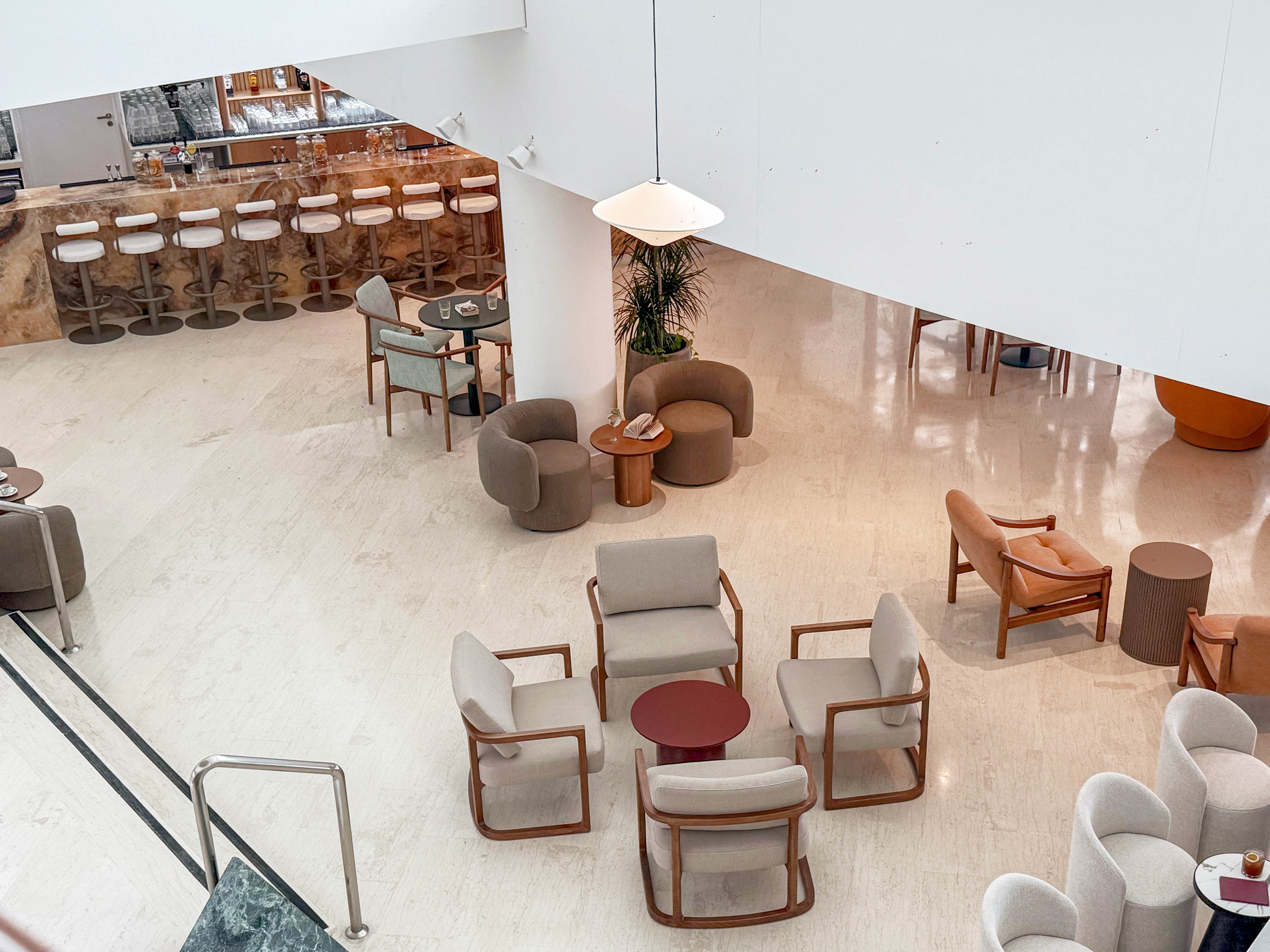 A contemporary lounge space features light-colored armchairs and small round tables arranged across a polished floor. In the background, a long bar counter with high stools lines the wall, suggesting a hotel or upscale lobby setting. The design emphasizes clean lines, neutral tones, and soft curves, creating a calm and inviting atmosphere. Large white walls and ample open space contribute to the airy, minimalist aesthetic.