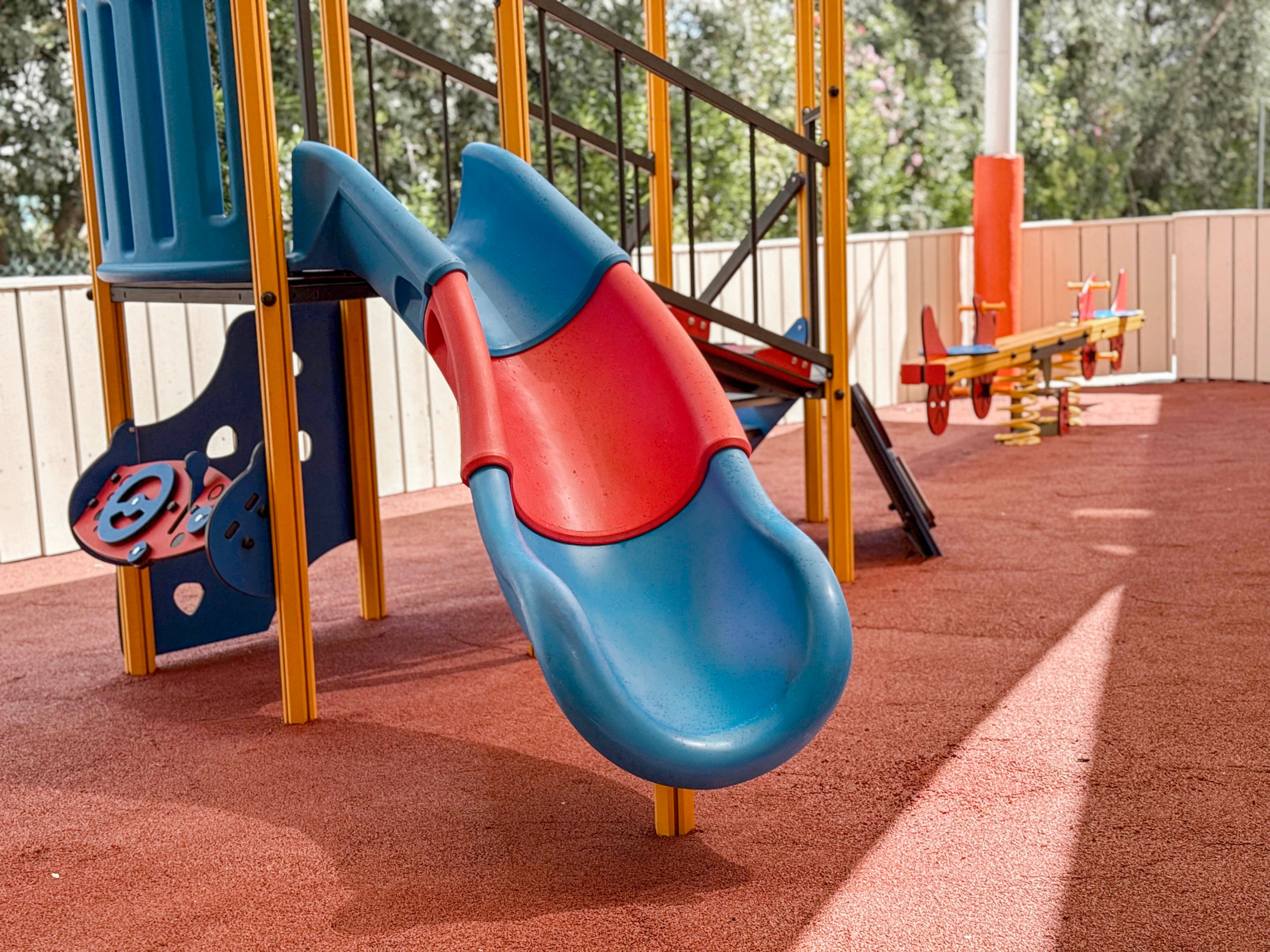 A young child is captured from behind sliding down a bright blue slide at an outdoor playground. The playground features red rubber flooring and yellow support structures, suggesting a safe and modern play area. Sunlight illuminates the scene, highlighting the child’s patterned sweater and the vivid primary colors of the equipment. Additional play structures are visible in the background, adding depth and context to the recreational setting.