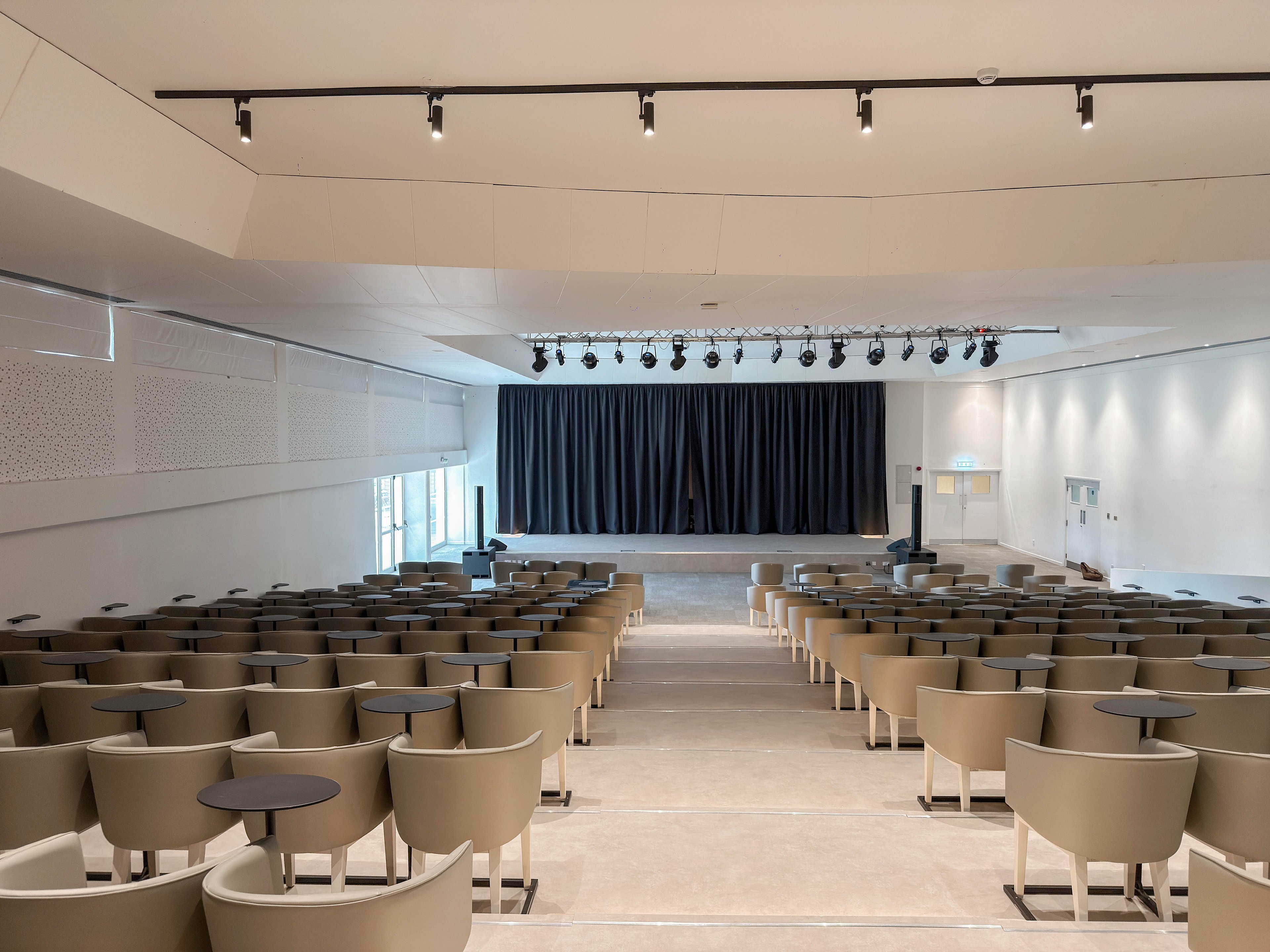 A modern auditorium features rows of beige armchairs facing a black-curtained stage. Small round tables are placed between the seats, suggesting a conference or lecture setting. The space is brightly lit with ceiling track lights and natural light from side windows. Clean white walls and minimalist design elements create a contemporary, professional atmosphere.