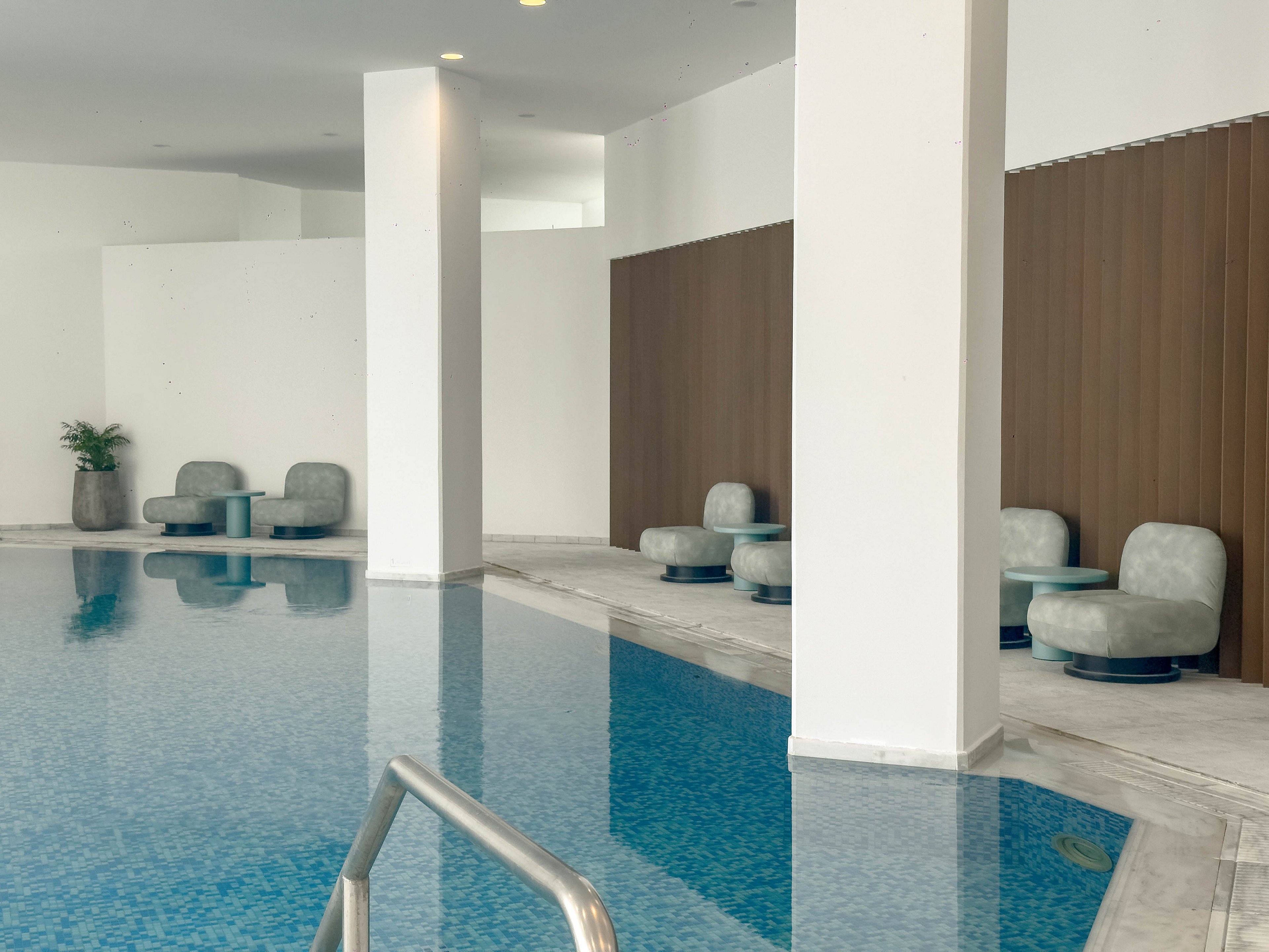 A serene indoor swimming pool features calm blue water and clean architectural lines. Soft upholstered lounge chairs with small side tables are arranged in alcoves along the poolside. Tall white columns and wood-paneled walls create a minimalist, contemporary atmosphere. A metal handrail leads into the pool, and a potted plant adds a touch of greenery to the tranquil setting.