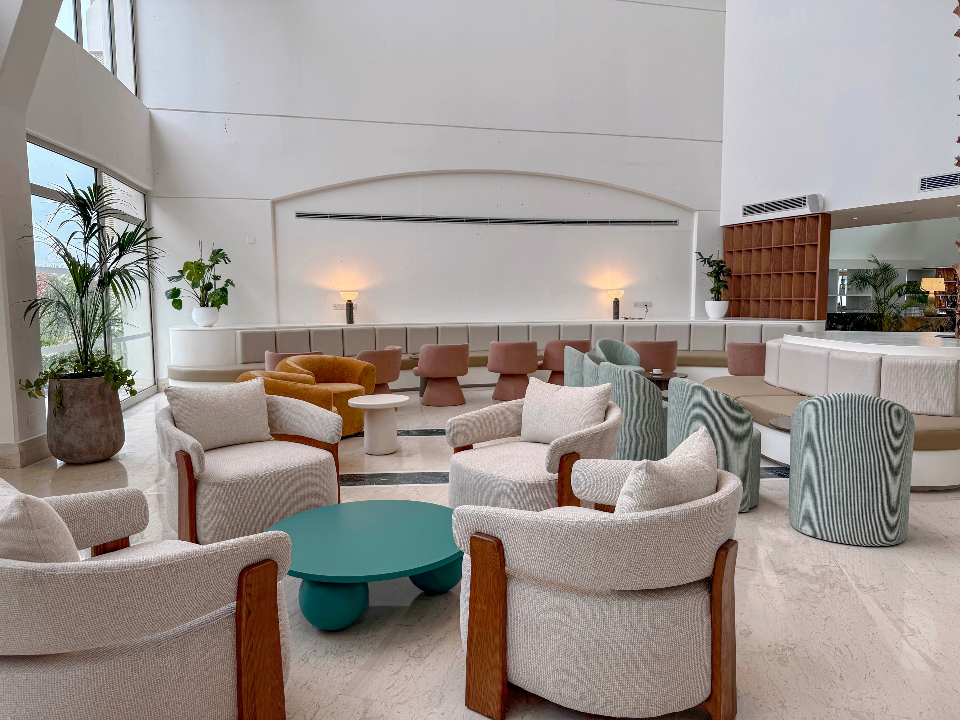 A contemporary hotel lobby lounge features clusters of cushioned armchairs and ottomans arranged around a teal coffee table. Neutral-toned seating in cream and beige is accented by a mustard yellow chair and a pale green ottoman. The space has high white walls, large windows, and potted plants adding greenery. Soft wall lighting and clean architectural lines create a calm, welcoming atmosphere suitable for relaxing or informal meetings.