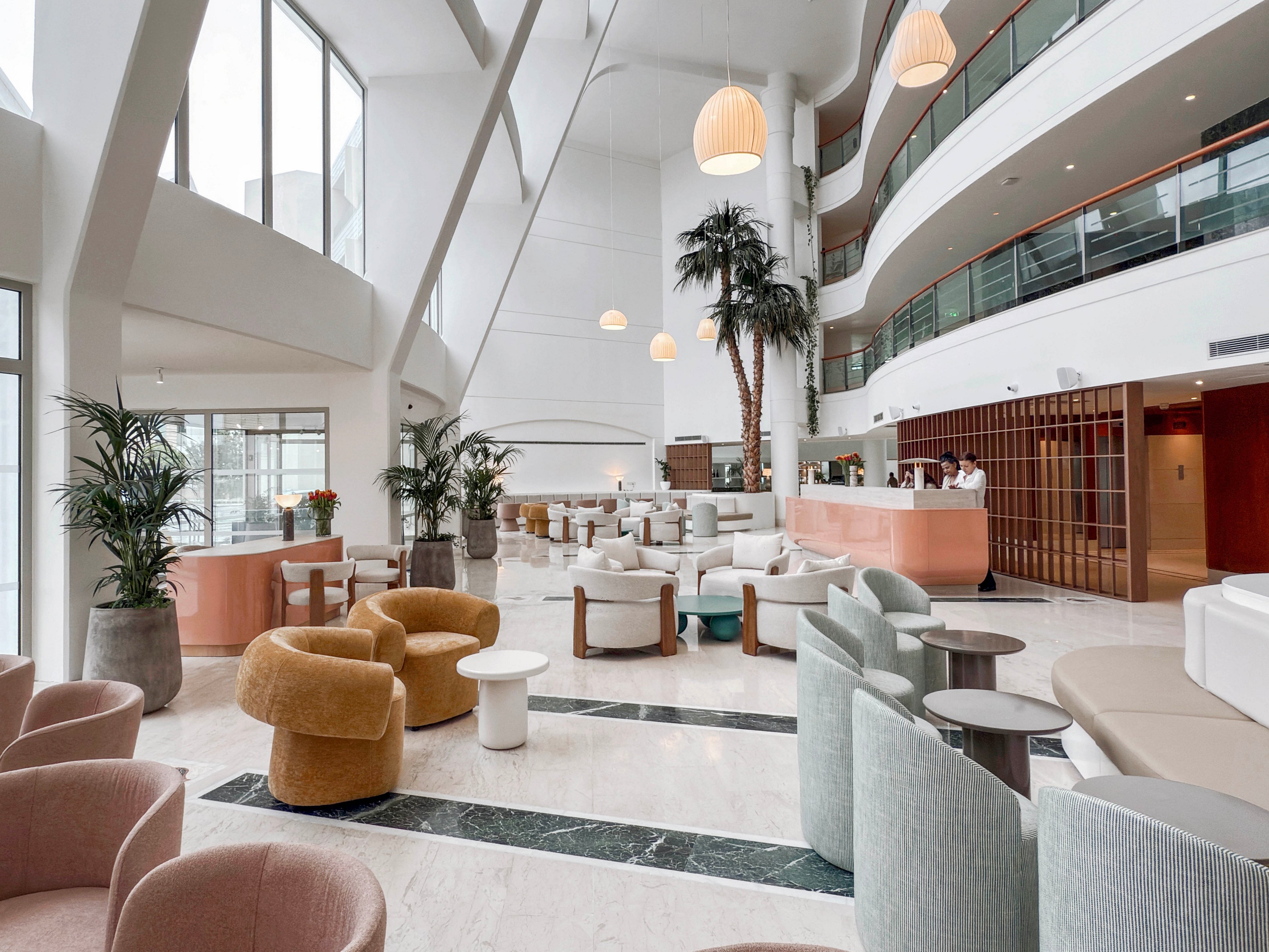Spacious hotel lobby interior featuring contemporary armchairs and small round tables. The setting includes high ceilings, large windows, and curved architectural lines creating an airy atmosphere. Soft pastel colors, indoor palm trees, and pendant lights contribute to a relaxed, upscale mood. A reception desk with staff is visible in the background near upper-level balconies.