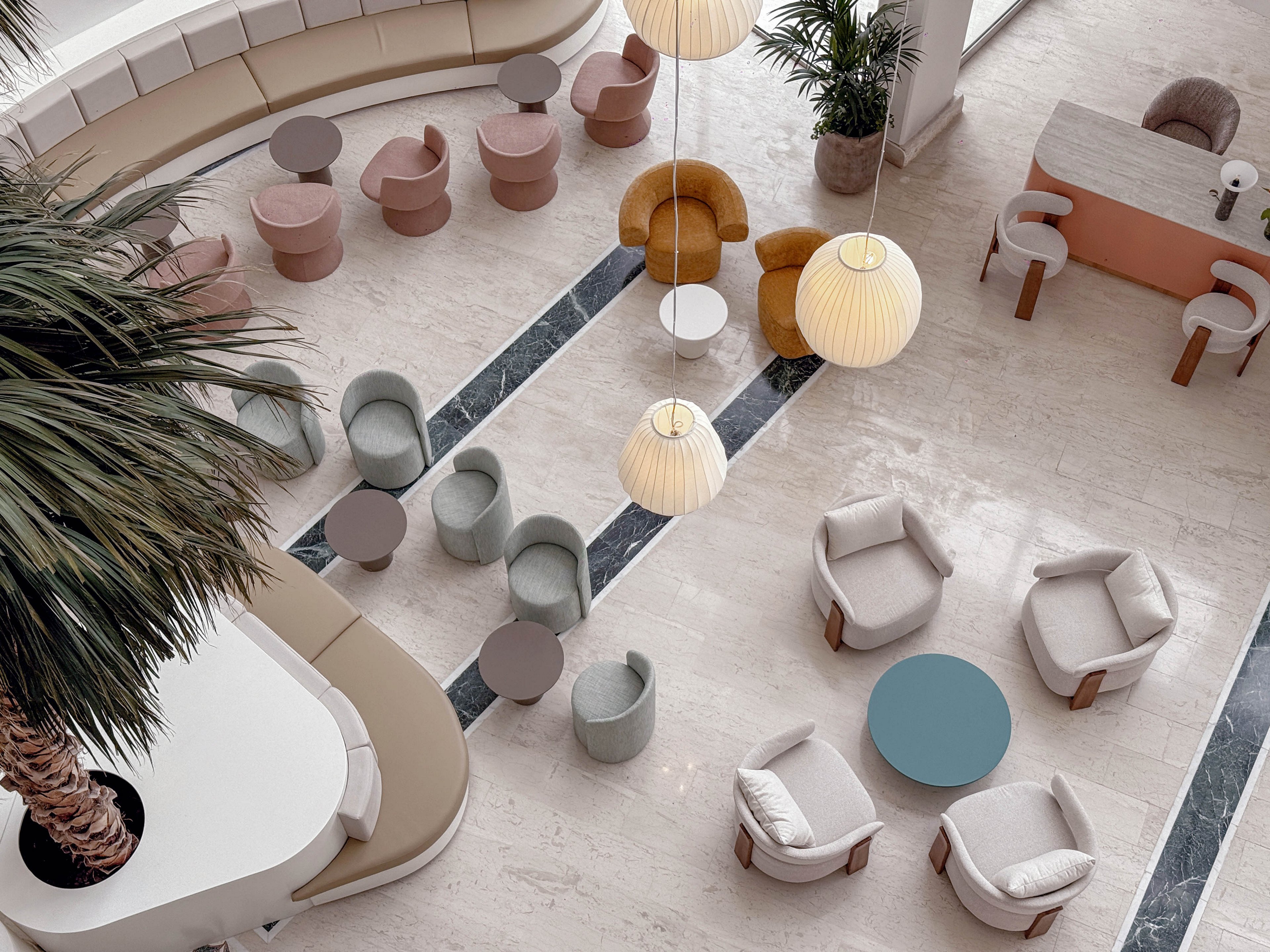 An overhead view shows a contemporary lounge area with curved banquettes, round tables, and cushioned chairs arranged on a light stone floor. Soft pastel tones of beige, blush, and muted teal create a calm, minimalist atmosphere. Large pendant lights hang above the seating, while a tall indoor palm tree adds a touch of greenery. A small bar or reception counter is visible in the background, enhancing the sense of a stylish social or hospitality space.