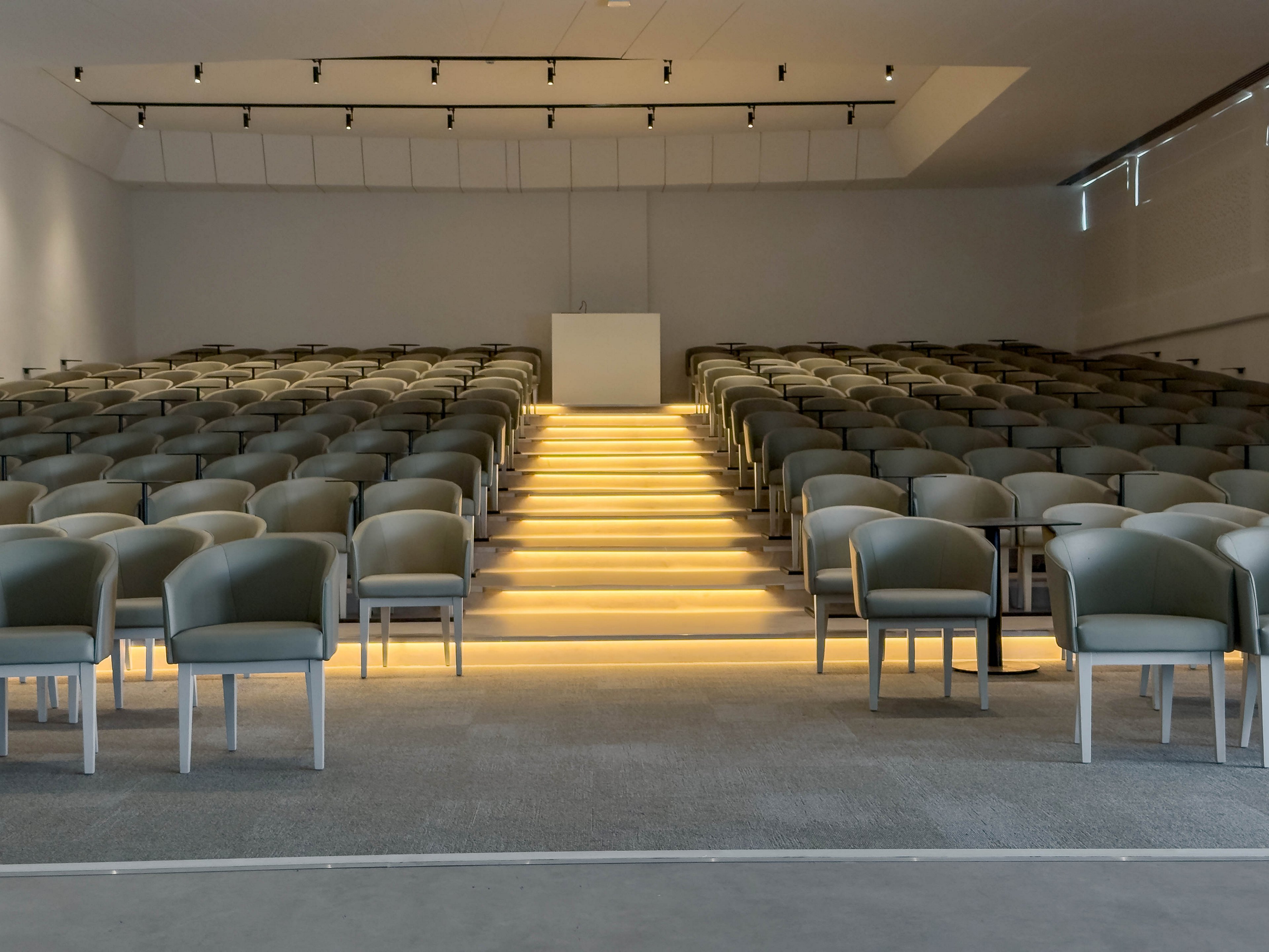 A contemporary auditorium features rows of empty cushioned chairs facing a central podium. Warmly lit steps lead up through the middle of the seating, creating a focal pathway to the front of the room. The space is minimalist, with neutral colors and clean architectural lines. Ceiling track lights and subtle wall lighting enhance the calm, professional atmosphere.