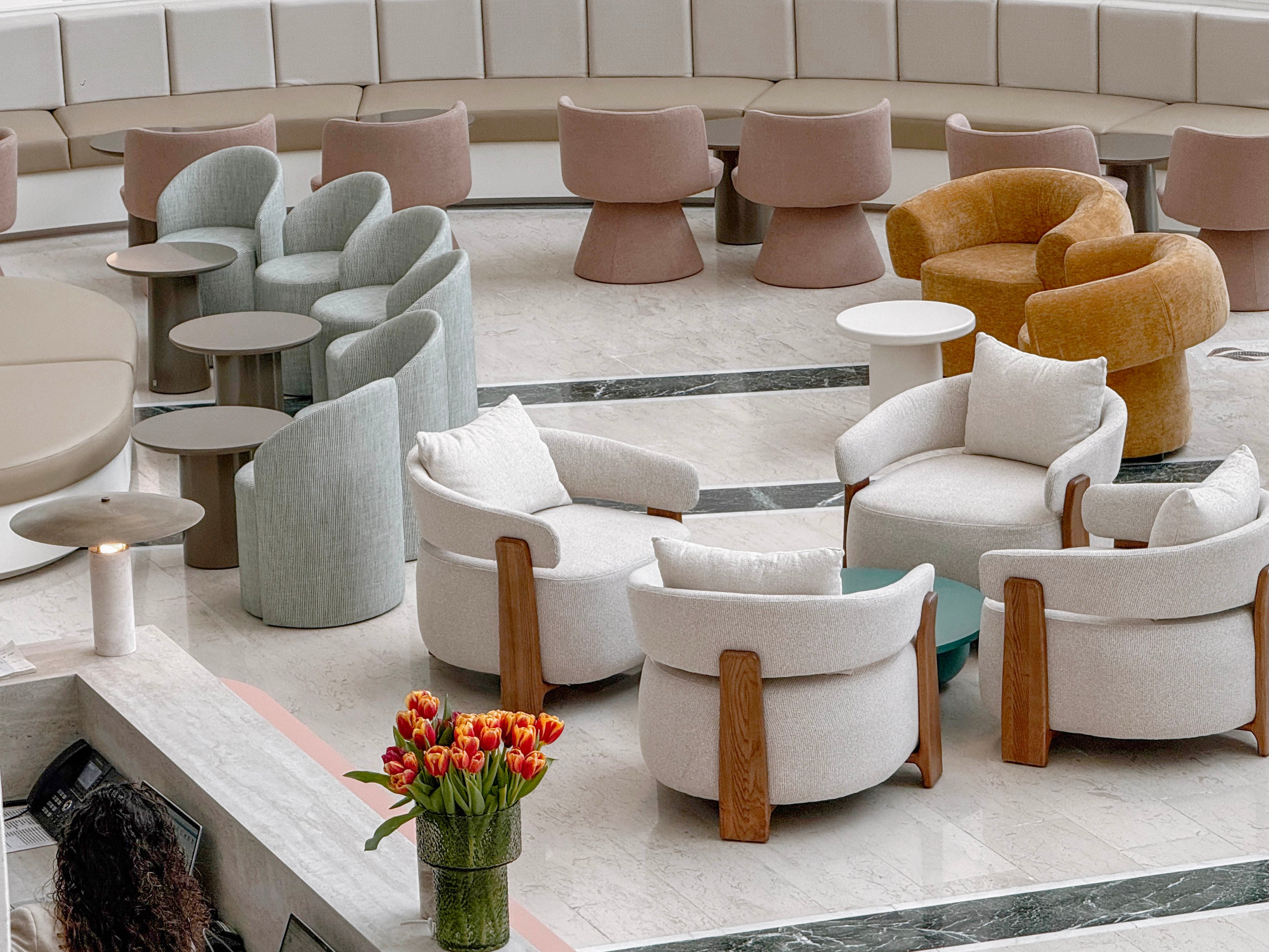 A collection of modern upholstered lounge chairs and small tables is arranged in a spacious indoor atrium. Soft pastel and neutral tones dominate the seating, including white, beige, light blue, and mustard yellow chairs. The marble or stone floor features dark inlaid lines that guide the eye through the space. A vase of red and orange tulips adds a bright accent near the seating area.