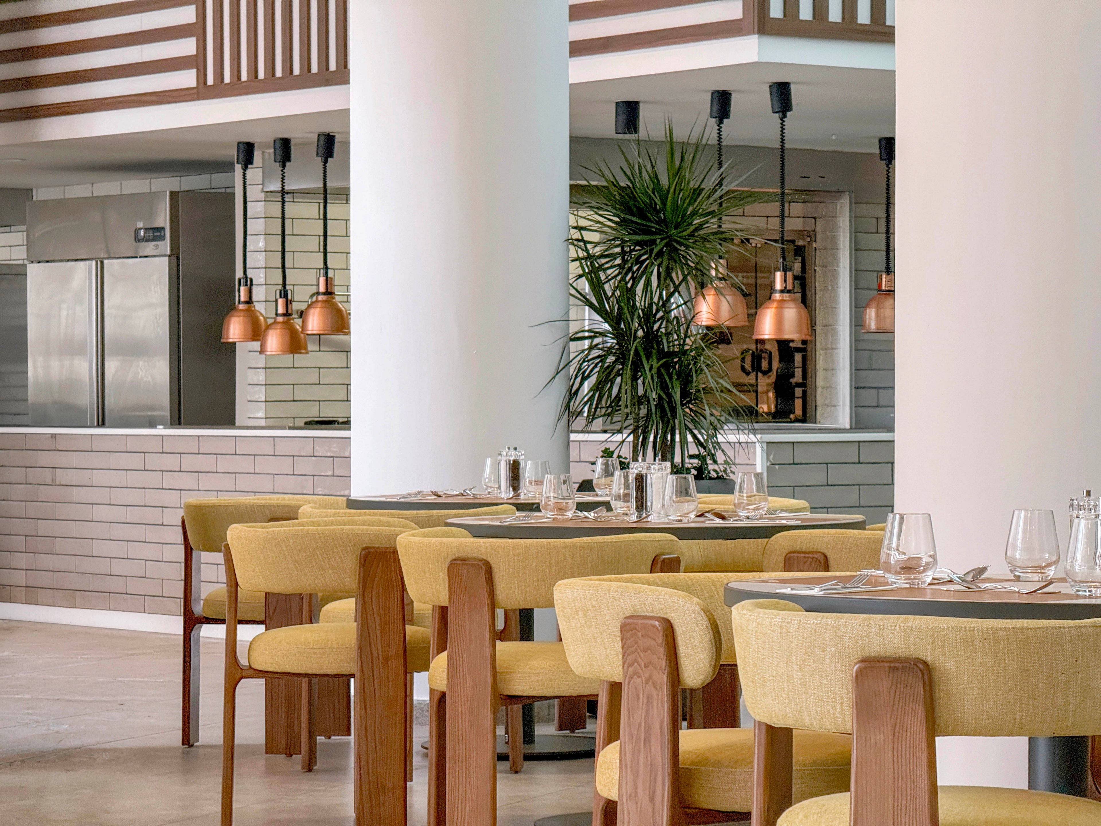A contemporary restaurant dining area features yellow upholstered chairs and wooden tables neatly set with glassware and cutlery. Large white columns divide the space, leading the eye toward an open kitchen with stainless steel appliances and white subway tiles. Warm copper pendant lights hang above the counter, adding a soft glow to the neutral palette. A tall indoor plant introduces greenery, enhancing the relaxed and inviting atmosphere.