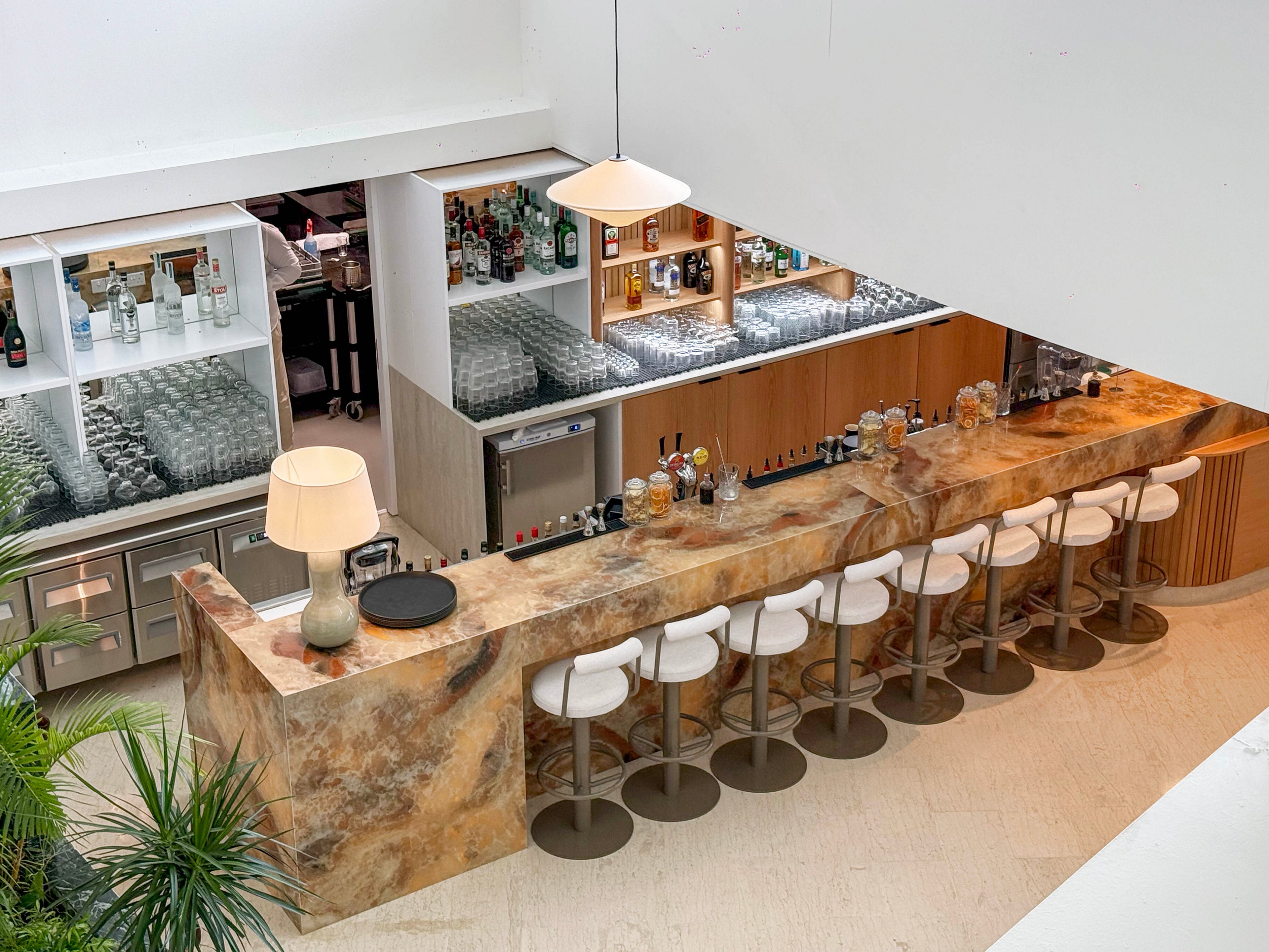 An elegant marble-topped bar is seen from above, lined with a row of white cushioned stools. The back bar features neatly arranged glassware and shelves stocked with various bottles of spirits. A warm table lamp and small decorative items sit on the counter, adding a cozy touch to the sleek, modern design. Green plants in the foreground soften the clean, minimalist interior.