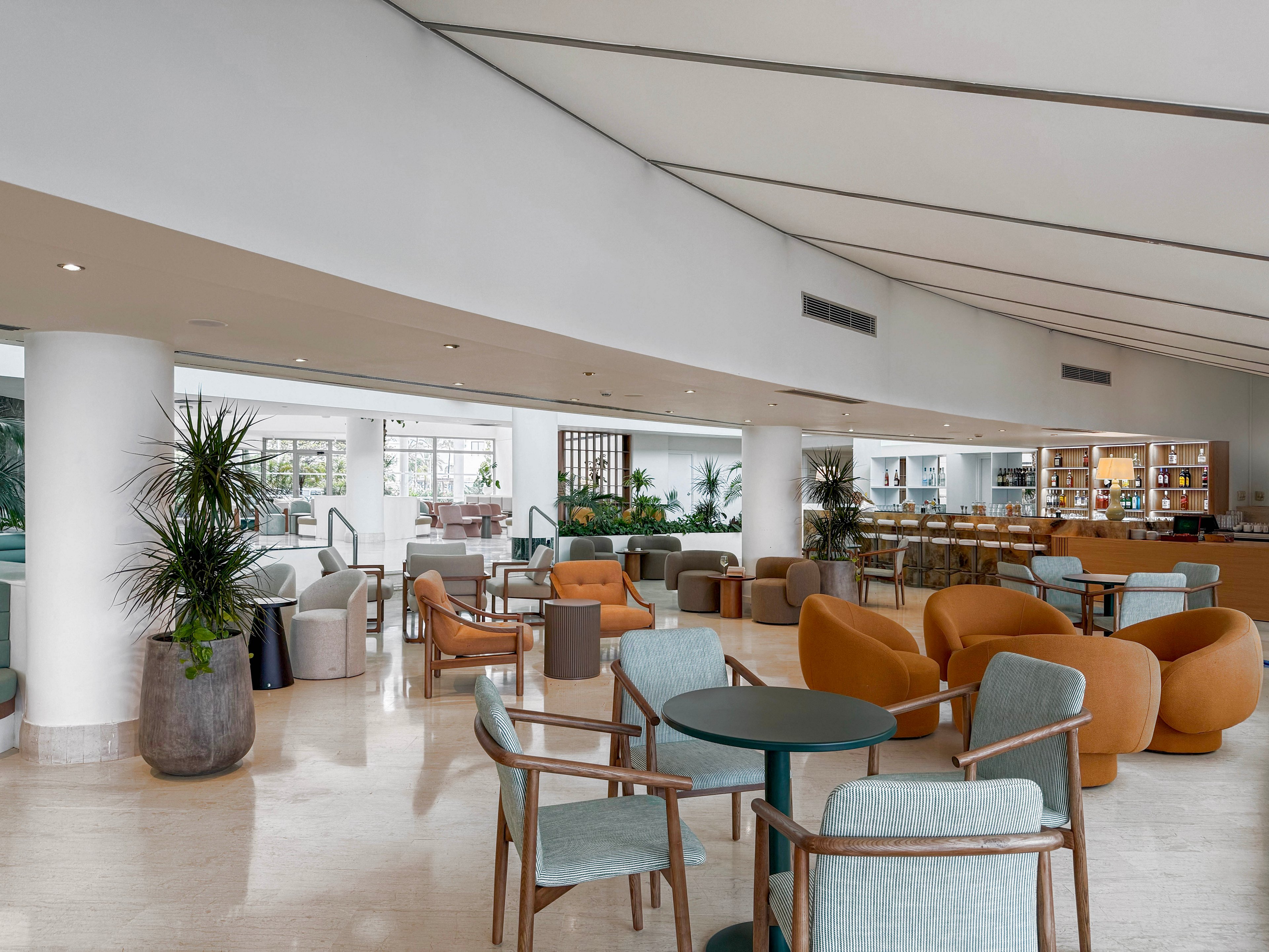 A spacious hotel lounge features modern armchairs and small round tables arranged across a polished floor. Soft blue and warm tan seating create a relaxed yet sophisticated atmosphere. Large windows in the background bring in natural light, while potted plants add greenery throughout the space. A bar area with shelves and bottles is visible at the back, enhancing the social and hospitality setting.