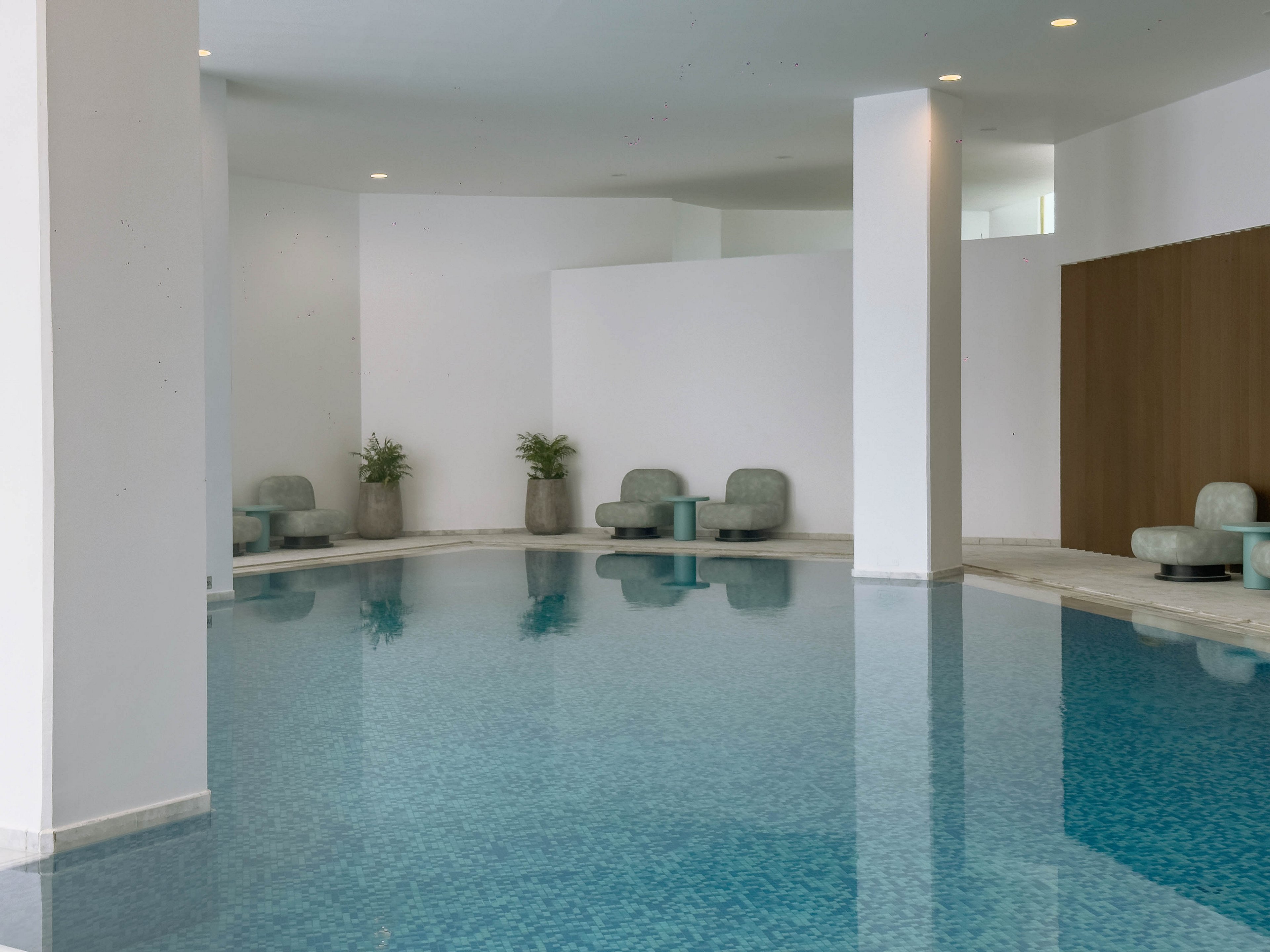 A serene indoor swimming pool is surrounded by clean white walls and large structural columns. Minimalist cushioned lounge chairs and small tables are arranged along the poolside, creating a quiet relaxation area. Potted plants add a touch of greenery to the otherwise neutral, modern space. Soft lighting and the blue water surface contribute to a calm, spa-like atmosphere.