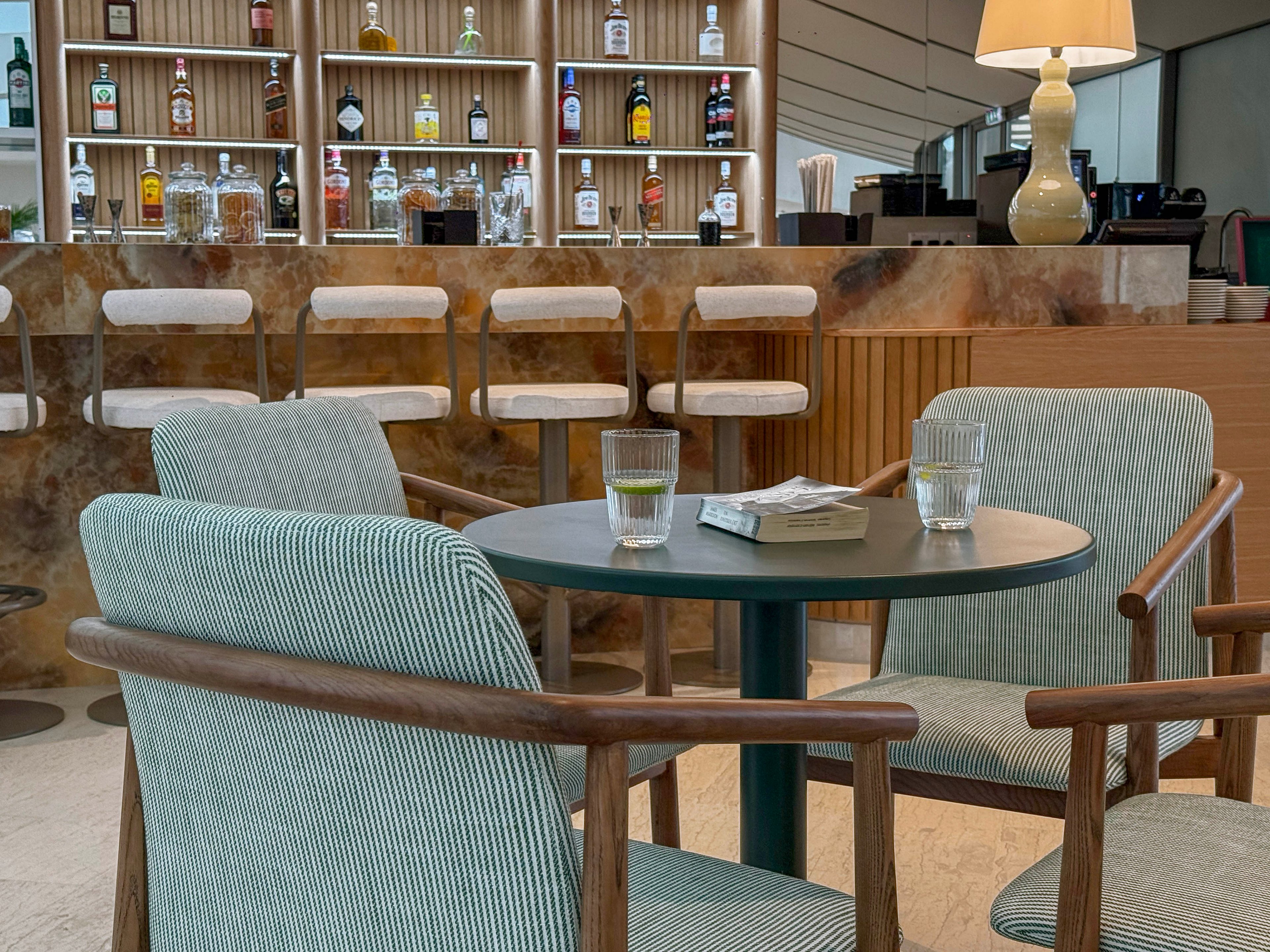 A contemporary lounge bar features a round table with two upholstered armchairs in the foreground. Two glasses of water and a closed book rest on the dark tabletop, suggesting a quiet break or meeting. In the background, a well-stocked bar displays various bottles and glassware on illuminated shelves. Neutral tones, soft fabrics, and warm lighting create a calm, sophisticated atmosphere.