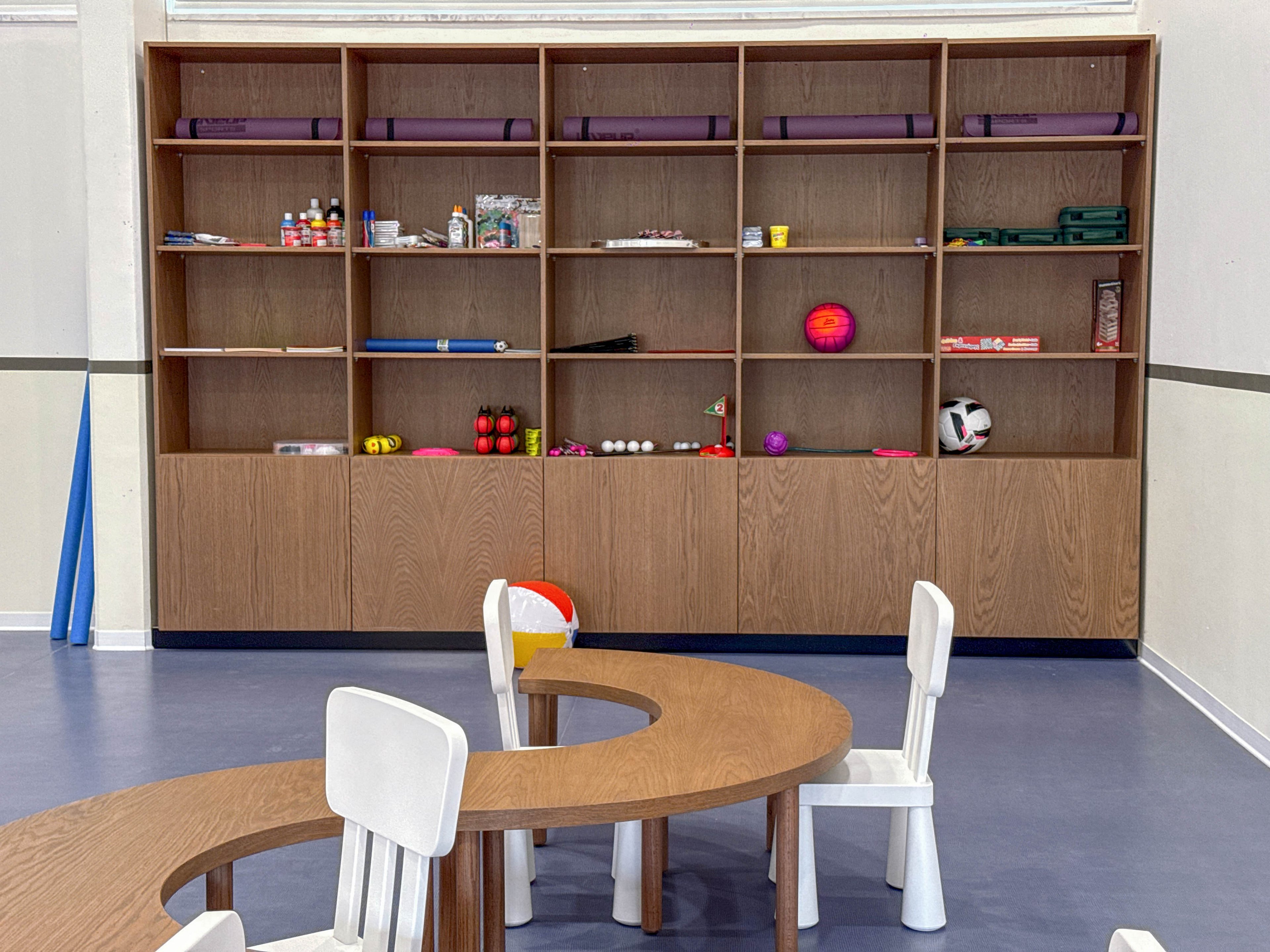 A bright playroom features a large wooden shelving unit filled with toys and supplies. In front of the shelves is a curved wooden table surrounded by small white chairs, suggesting a space for children’s activities. Various balls, blocks, and craft materials are neatly arranged on the shelves, adding pops of color against the neutral walls and floor. The room appears organized and inviting, suitable for learning, play, or group activities.