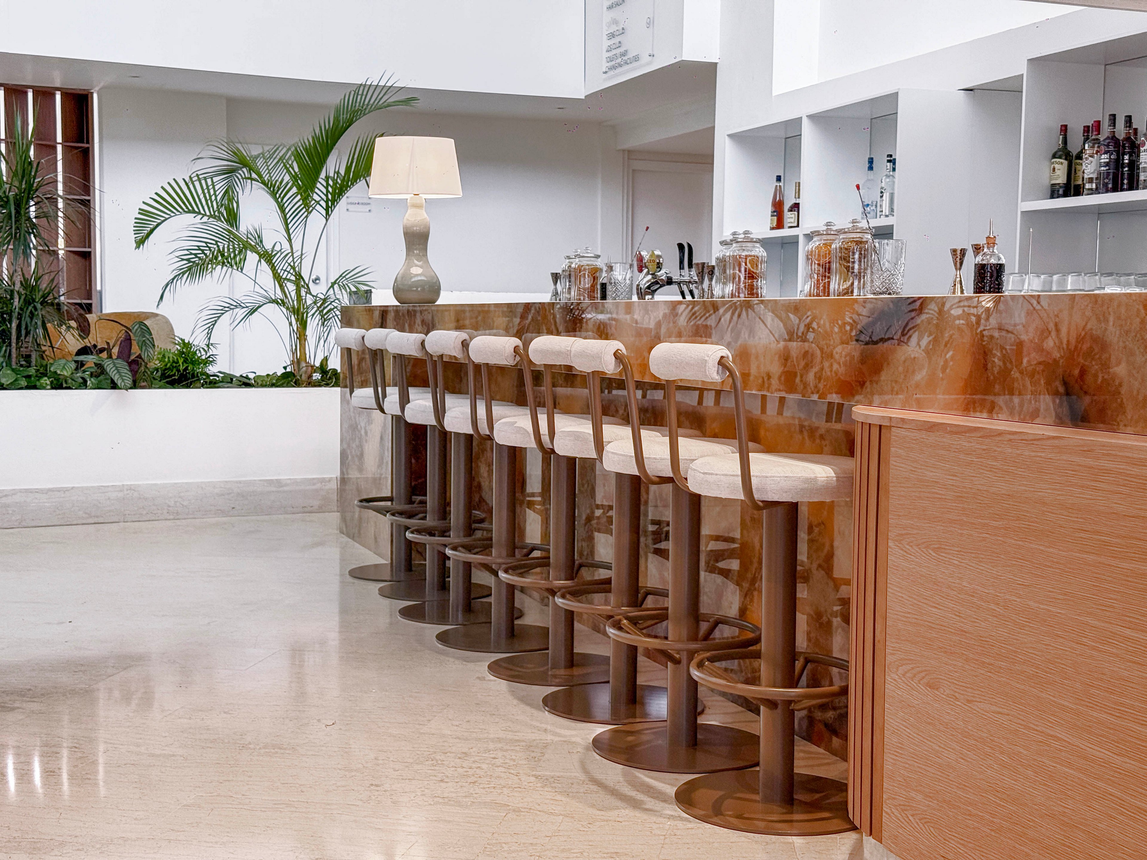 A sleek, contemporary bar features a polished marble counter with a row of empty bar stools. The setting appears to be a bright hotel or restaurant interior with light-colored flooring and minimalist decor. Bottles and glassware are neatly arranged on shelves behind the bar, while a table lamp adds a warm accent. Green plants soften the clean lines, creating a relaxed yet sophisticated atmosphere.