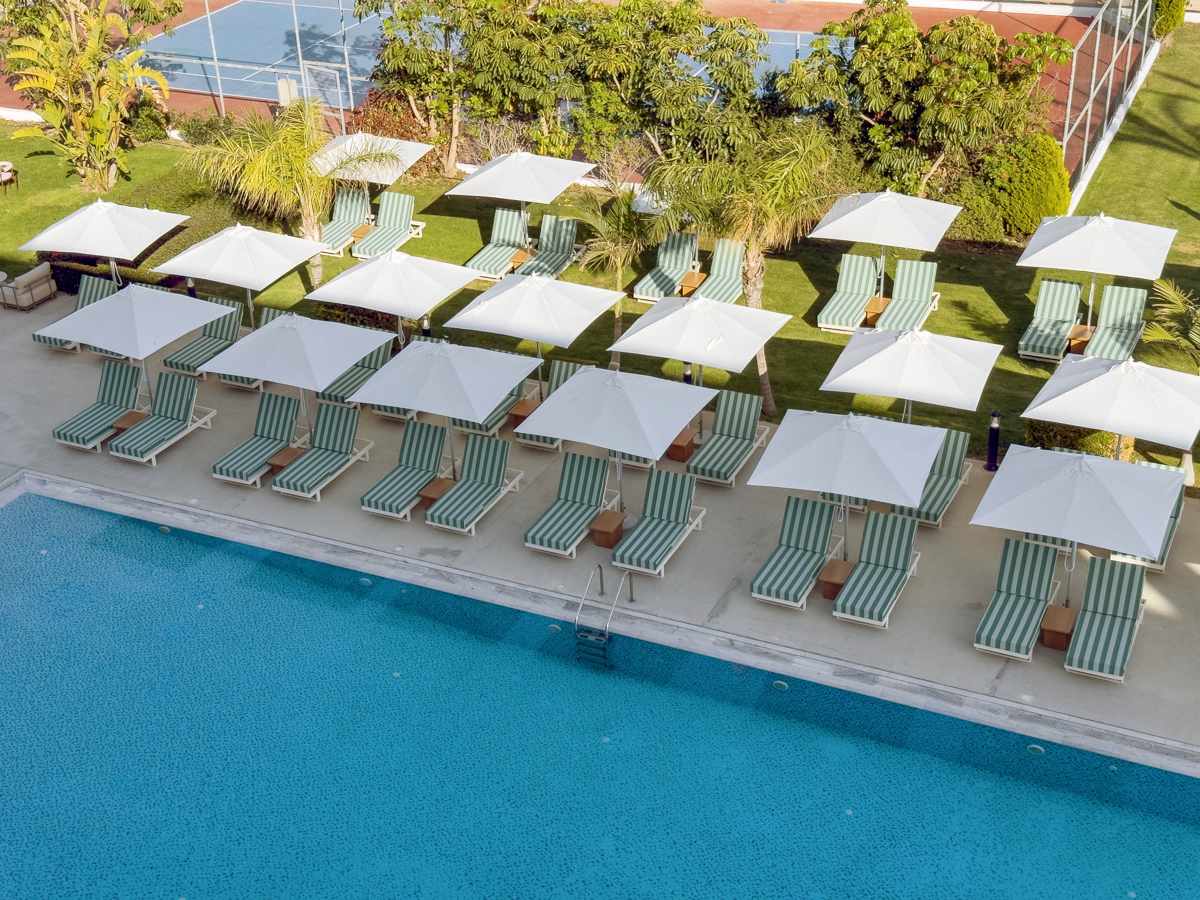 An aerial view shows a row of striped lounge chairs neatly arranged beside a clear blue swimming pool. White umbrellas provide shade over each lounger, creating a uniform and orderly pattern. The surrounding area includes green lawns, palm-like plants, and a tennis court in the background. The scene suggests a quiet, upscale resort environment ready for guests.