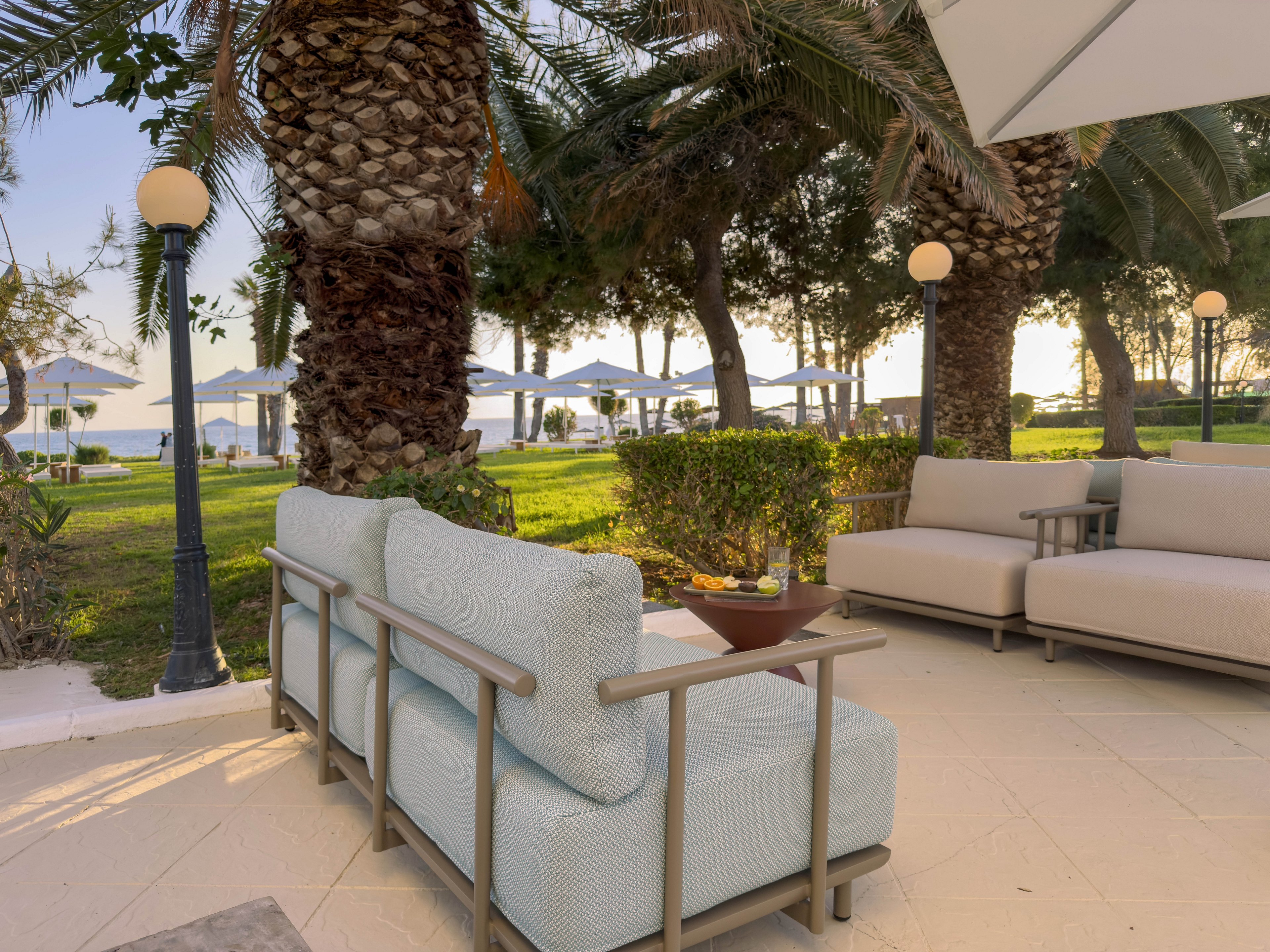 An inviting outdoor lounge area features cushioned patio sofas arranged on a tiled terrace. The seating overlooks a lush lawn with palm trees and trimmed bushes leading toward the water. White umbrellas and lounge chairs are visible in the background near the shoreline, suggesting a resort or hotel setting. Warm, soft lighting and calm surroundings create a relaxed, vacation-like atmosphere.