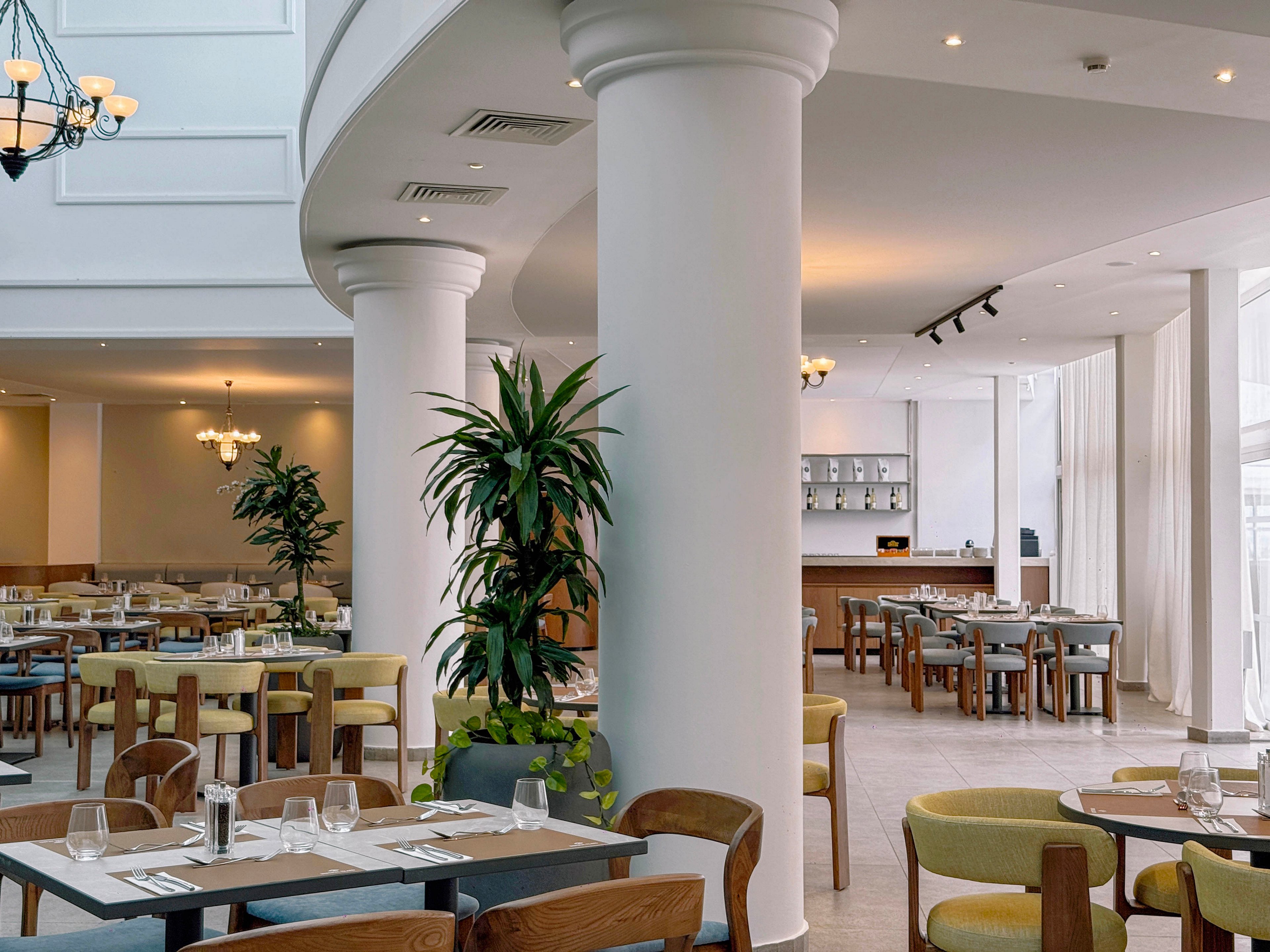 A spacious modern restaurant interior features large white columns and neatly arranged dining tables. Wooden chairs with light green and beige upholstery surround the tables, each set with glasses and cutlery. Tall potted plants add greenery to the bright space, which is illuminated by natural light from large windows and ceiling fixtures. In the background, a bar or service counter is visible, enhancing the contemporary hotel dining atmosphere.