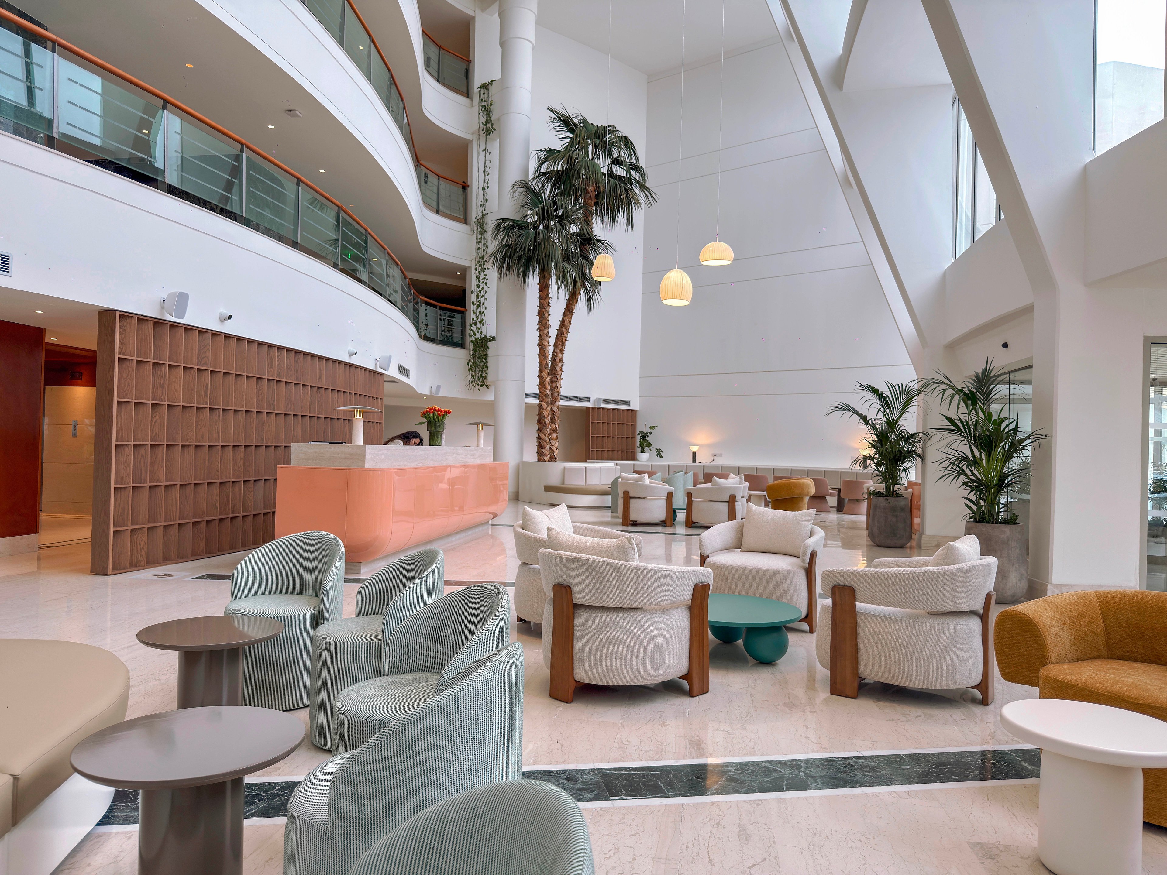 A spacious modern lobby features contemporary lounge seating arranged in small conversation areas. Soft pastel armchairs and round tables sit on a polished marble floor beneath high white walls and large windows. Tall indoor palm trees and potted plants add greenery to the bright, minimalist space. A curved reception desk and upper-level balconies emphasize the open, airy architectural design.