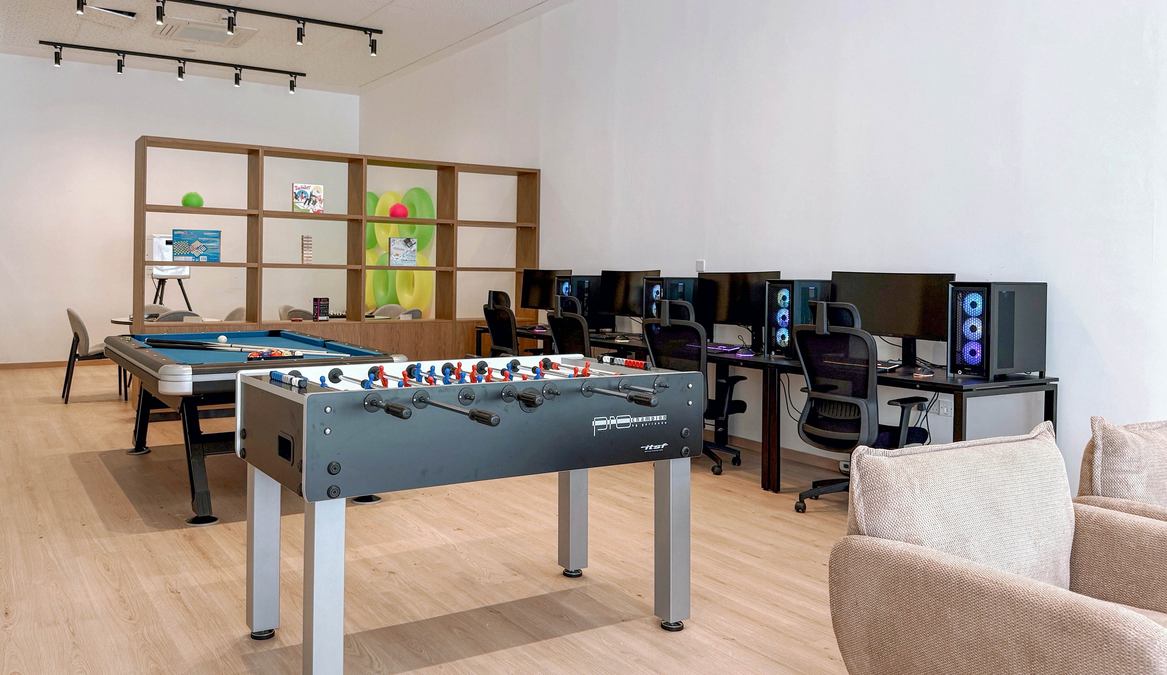A contemporary gaming lounge features a foosball table in the foreground and a row of desktop gaming computers along the wall. Each computer station includes a large monitor, keyboard, mouse, and tower with illuminated blue fans. In the background, a pool table and open shelving add to the recreational atmosphere. The room has light wooden flooring, white walls, and neutral-toned seating, creating a clean and inviting space for leisure and gaming.