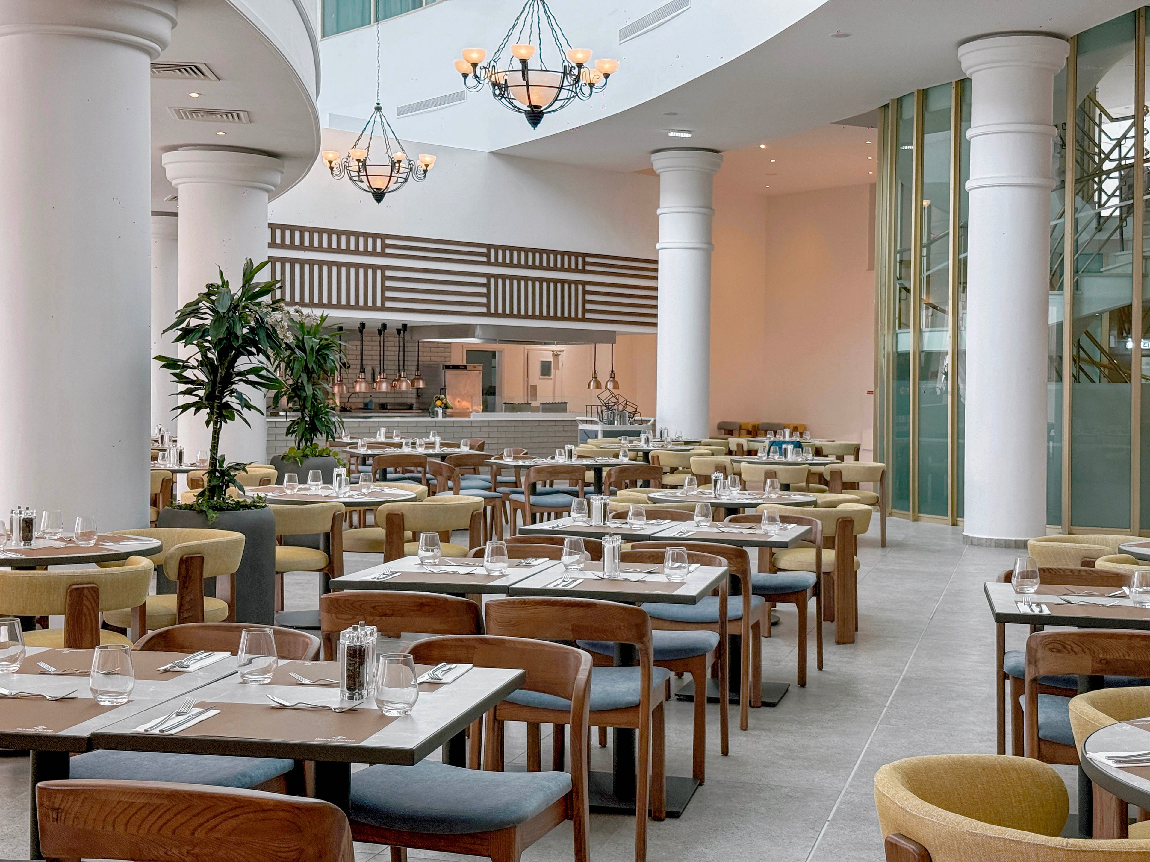 Contemporary restaurant interior featuring neatly arranged wooden tables and upholstered chairs. The setting includes high white columns, large windows, and an open kitchen area in the background. Table settings with glassware, cutlery, and napkins create a refined yet casual dining atmosphere. Soft neutral tones and natural light contribute to a clean, modern, and welcoming ambiance.
