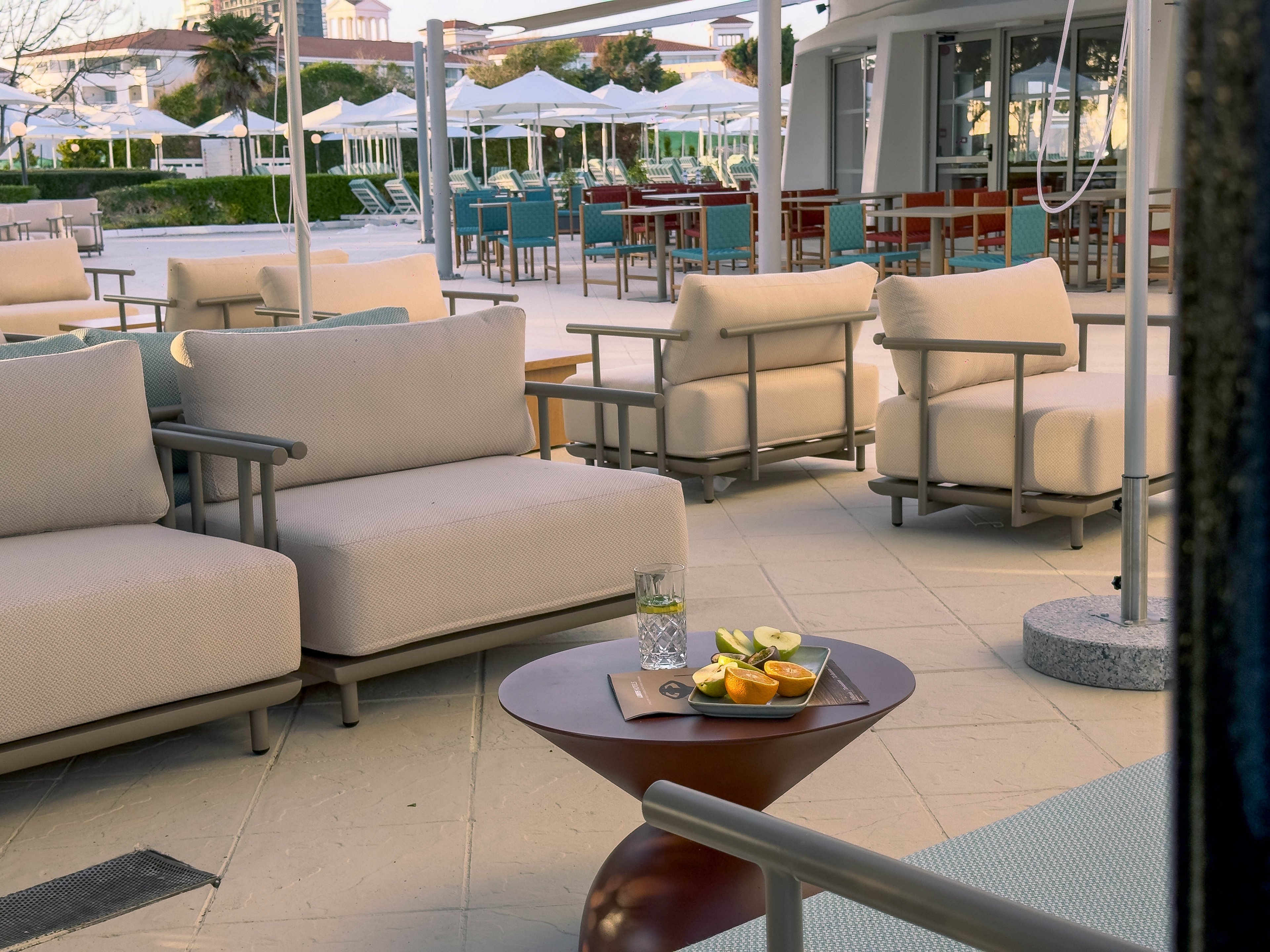 An outdoor lounge area features modern cushioned sofas and armchairs arranged on a tiled patio. In the foreground, a small round table holds a glass of water and a plate with sliced citrus fruits. The background shows additional seating and tables under umbrellas, suggesting a relaxed hospitality setting. Soft neutral tones and clean lines create a calm, contemporary atmosphere.