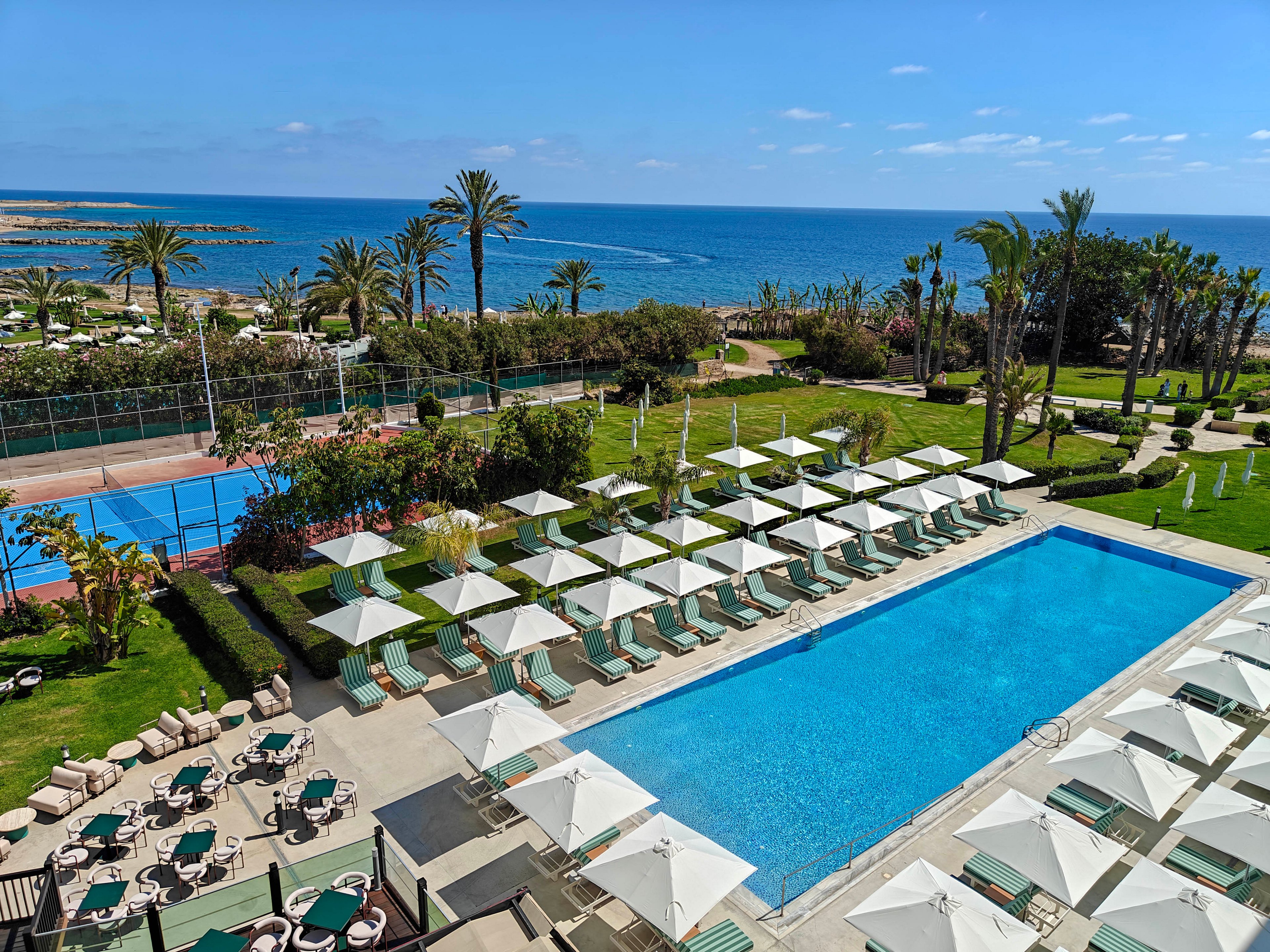 A large outdoor swimming pool is surrounded by rows of white sun umbrellas and lounge chairs at a coastal resort. The scene overlooks manicured lawns, palm trees, and a tennis court near the shoreline. Beyond the property, the blue ocean stretches to the horizon under a clear sky with a few scattered clouds. The overall atmosphere is calm, sunny, and inviting for vacation relaxation.