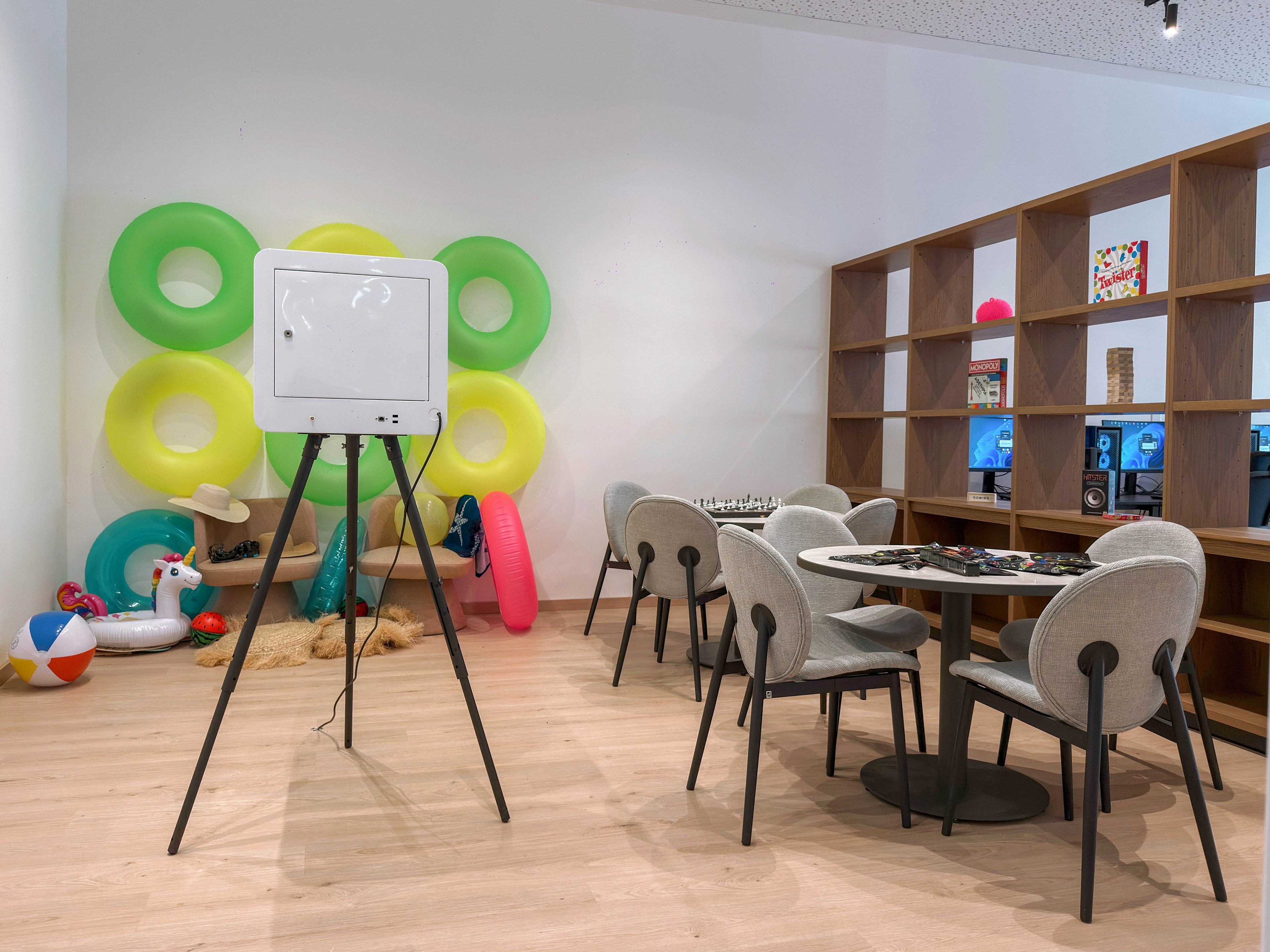 A contemporary lounge area features a small round table with chairs set up for board games and foosball. In the foreground, a white device on a tripod stands on the wooden floor. The back corner displays colorful pool inflatables, a beach ball, and a straw hat, adding a playful, summery touch. Open shelving along the wall holds books, games, and decorative items, creating a relaxed, social atmosphere.