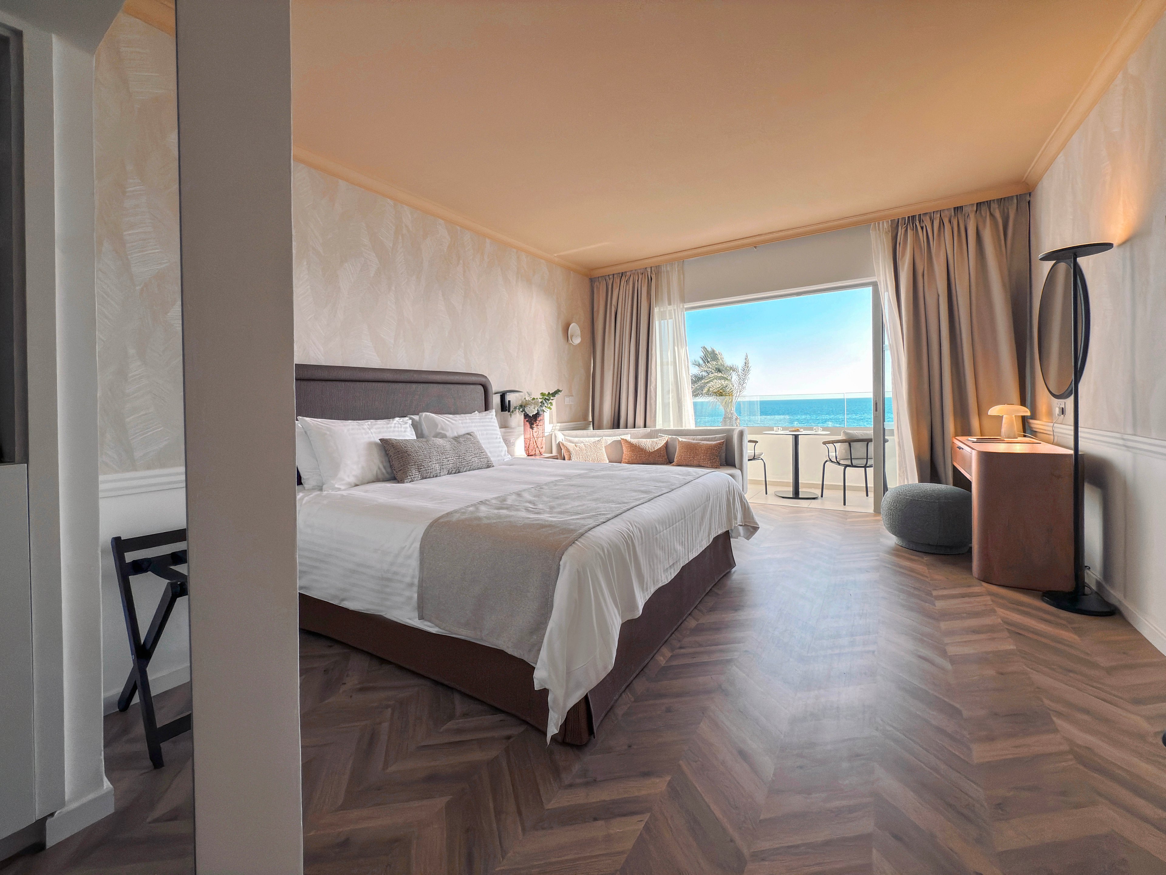A contemporary hotel bedroom features a neatly made double bed with neutral-toned linens and accent pillows. The room opens onto a balcony with chairs and a table overlooking a bright blue ocean. Warm lighting, wooden herringbone flooring, and minimalist furniture create a calm, inviting atmosphere. Floor-to-ceiling curtains frame the large window, enhancing the coastal view and airy feel of the space.