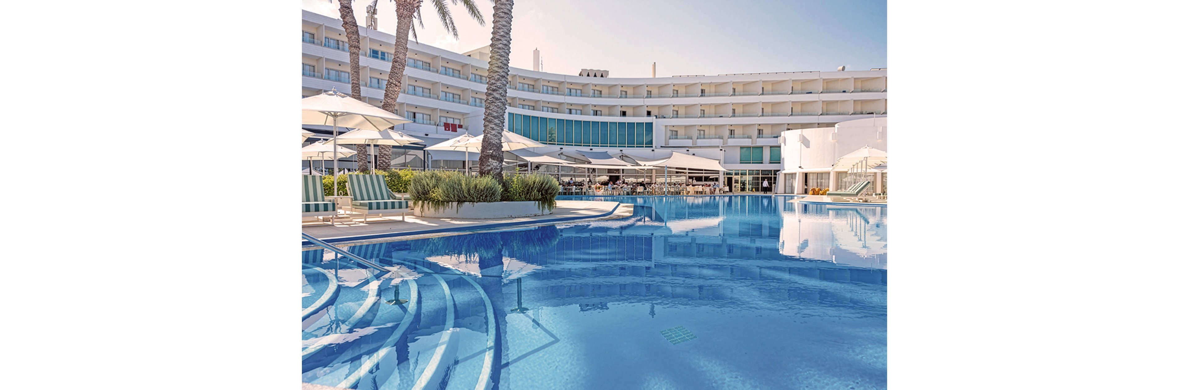 A large outdoor swimming pool reflects a modern multi-story hotel building under a clear sky. Sun loungers and umbrellas line the poolside terrace, creating a relaxed vacation atmosphere. Tall palm trees and low ornamental plants add a tropical touch to the scene. The overall setting suggests a sunny resort destination focused on leisure and comfort.