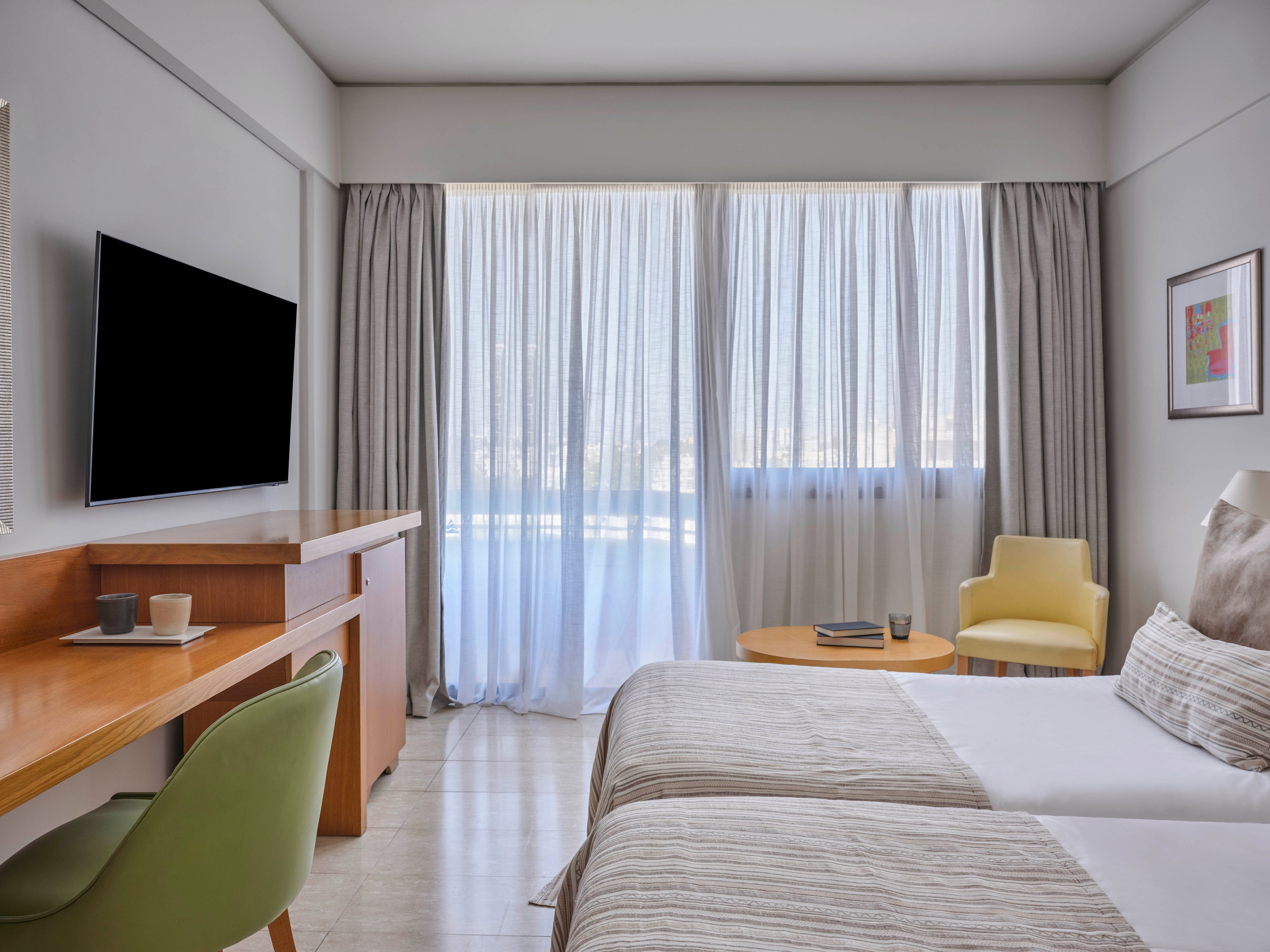 A contemporary hotel room features two neatly made beds with neutral bedding. Floor-to-ceiling windows with sheer and blackout curtains allow soft daylight to fill the space. A wall-mounted flat-screen TV faces a wooden desk with a green chair and a cup placed on top. In the corner, a yellow armchair and round wooden table with a glass and book create a cozy seating area, complemented by a framed artwork on the wall.