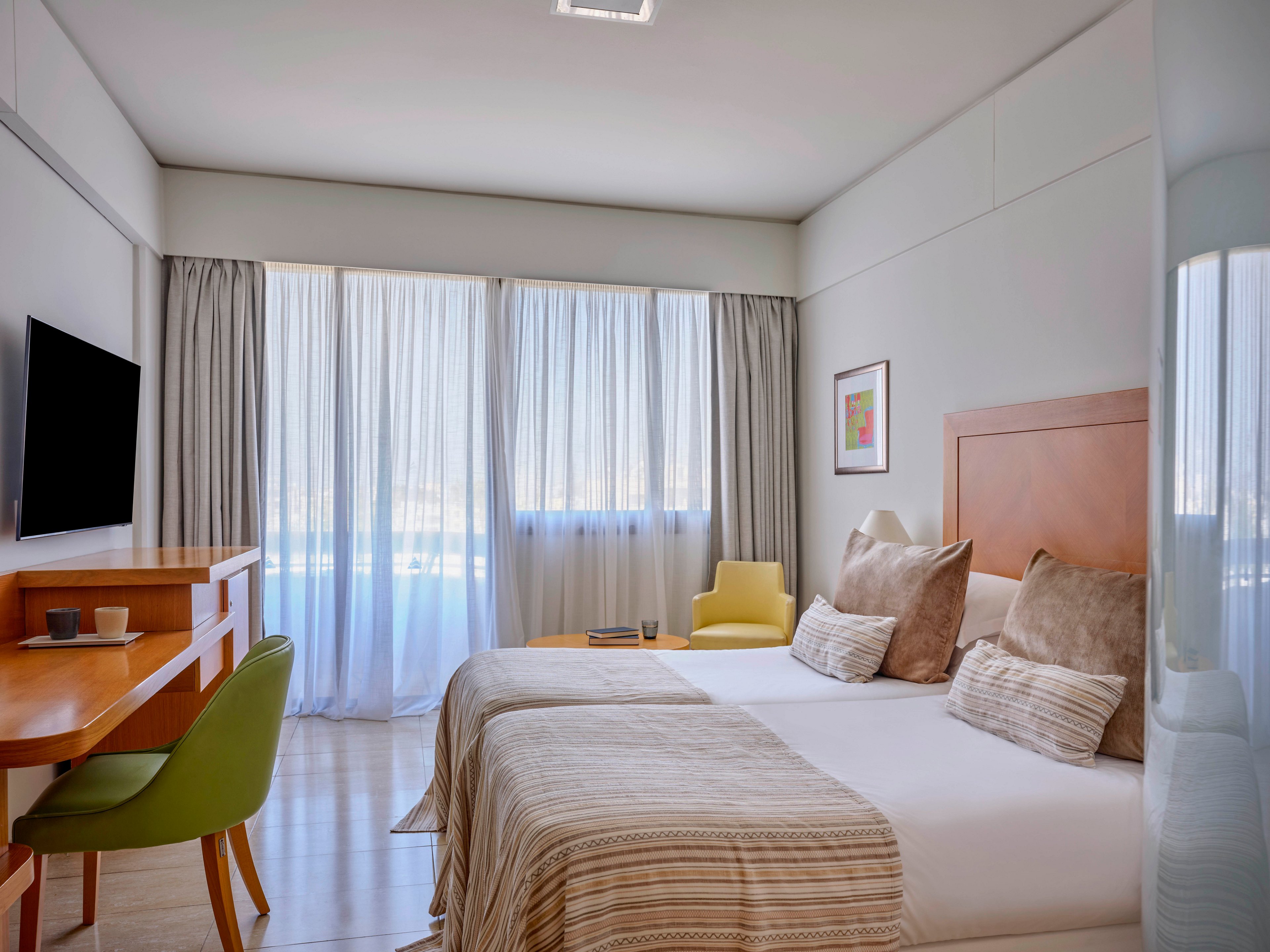 A contemporary hotel room features two neatly made twin beds with neutral bedding and multiple cushions. Natural light streams through large sheer curtains covering floor-to-ceiling windows, creating an airy atmosphere. A wooden desk with a green chair faces a wall-mounted television, while a yellow armchair sits near the window. Soft beige tones, clean lines, and minimal decor give the space a calm, inviting feel.
